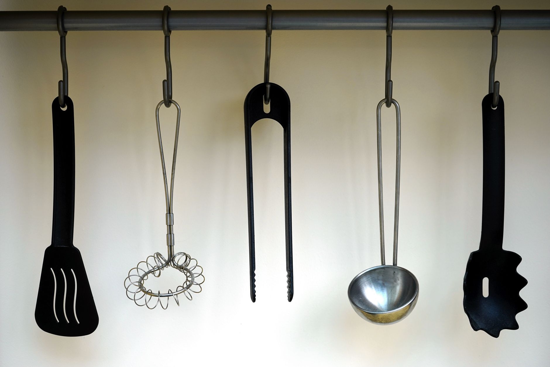 Espátula, batidor, pinzas y cucharón colgados de ganchos en una barra de pared de la cocina
