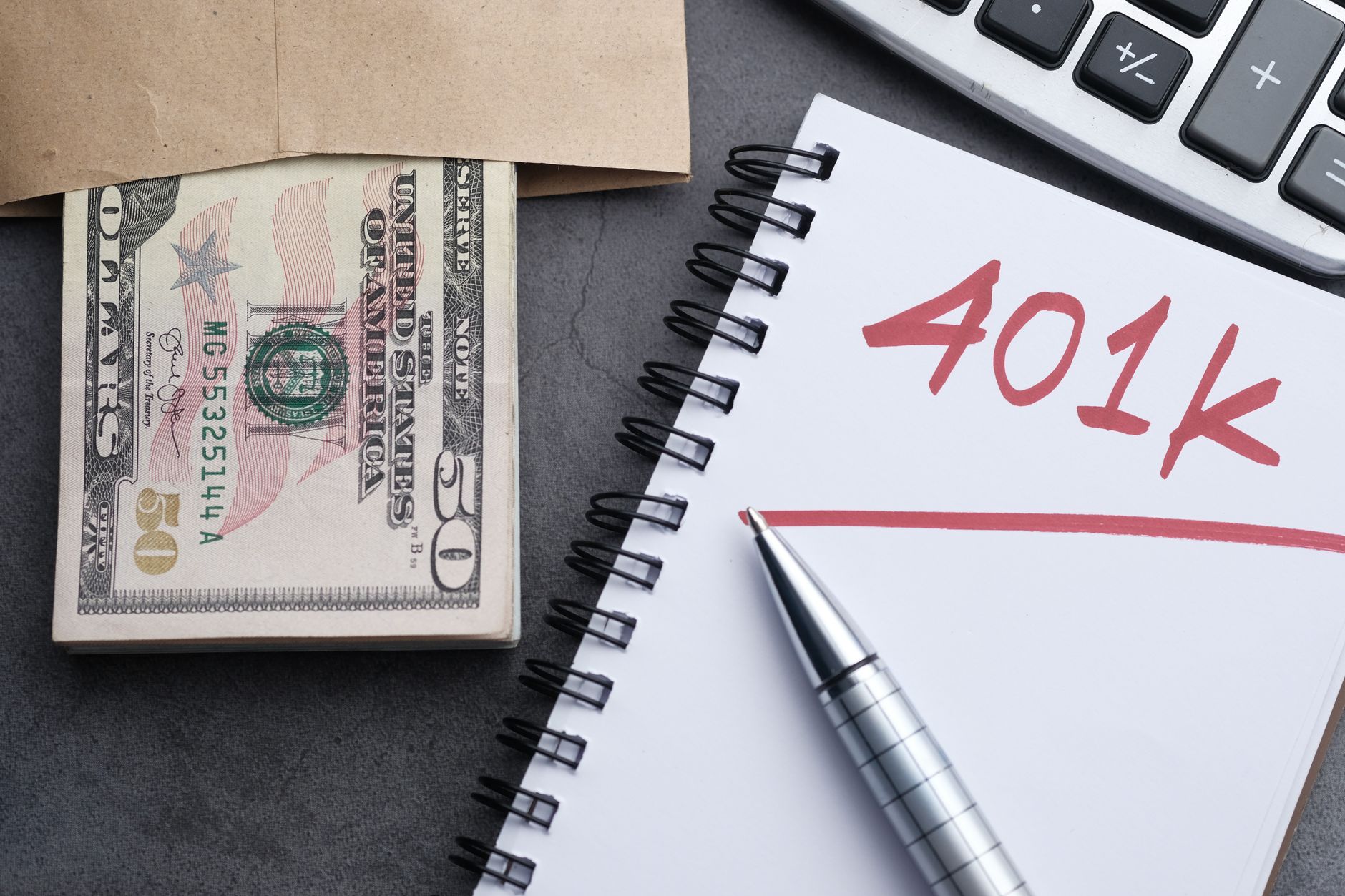 Libreta con la palabra 401k escrita a mano junto a un fajo de billetes en un sobre, un bolígrafo y una calculadora
