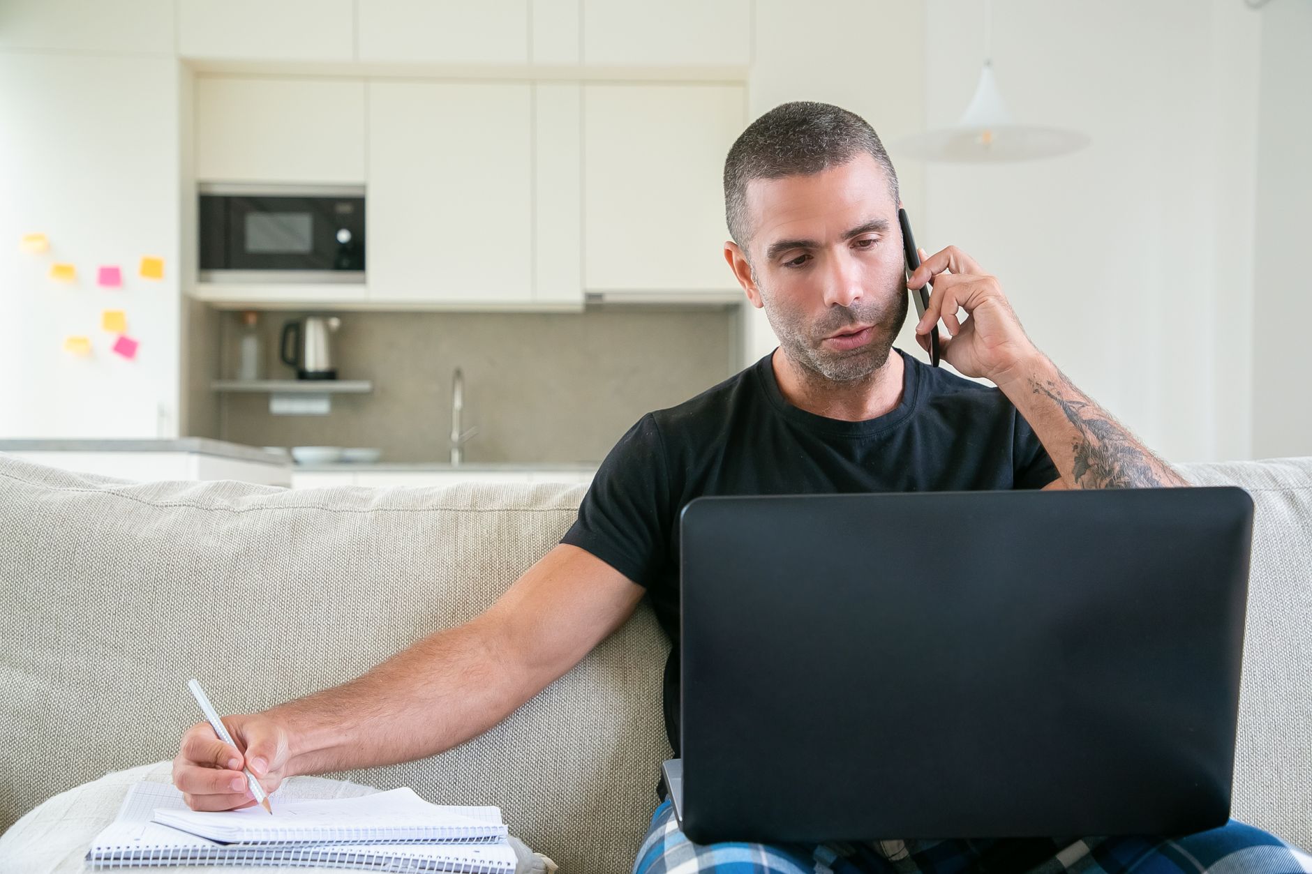 Hombre tomando notas en una libreta mientras habla por teléfono con la computadora abierta en casa