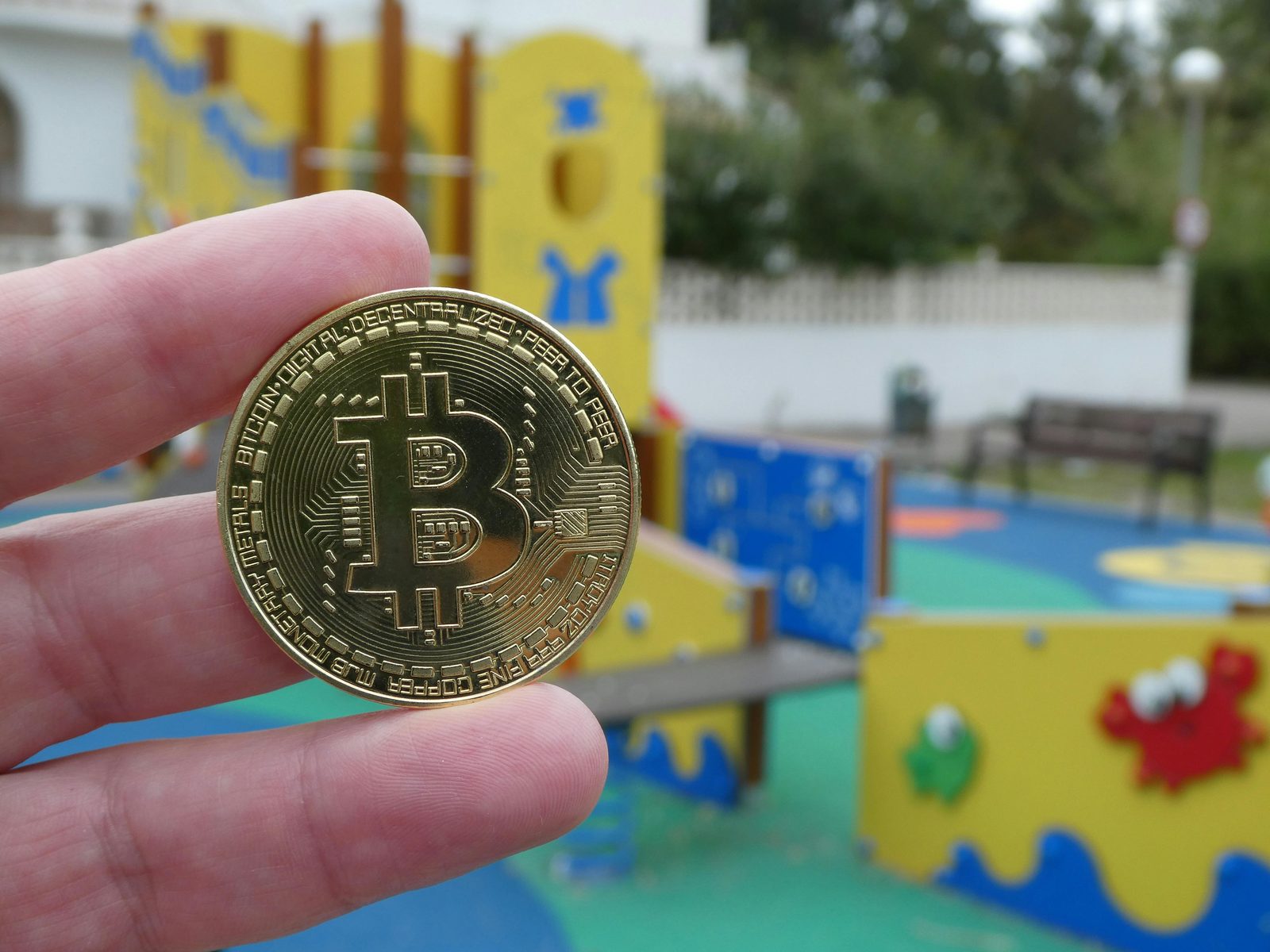Mano sosteniendo una moneda dorada de Bitcoin con el texto BITCOIN DIGITAL PEER TO PEER frente a un parque infantil.