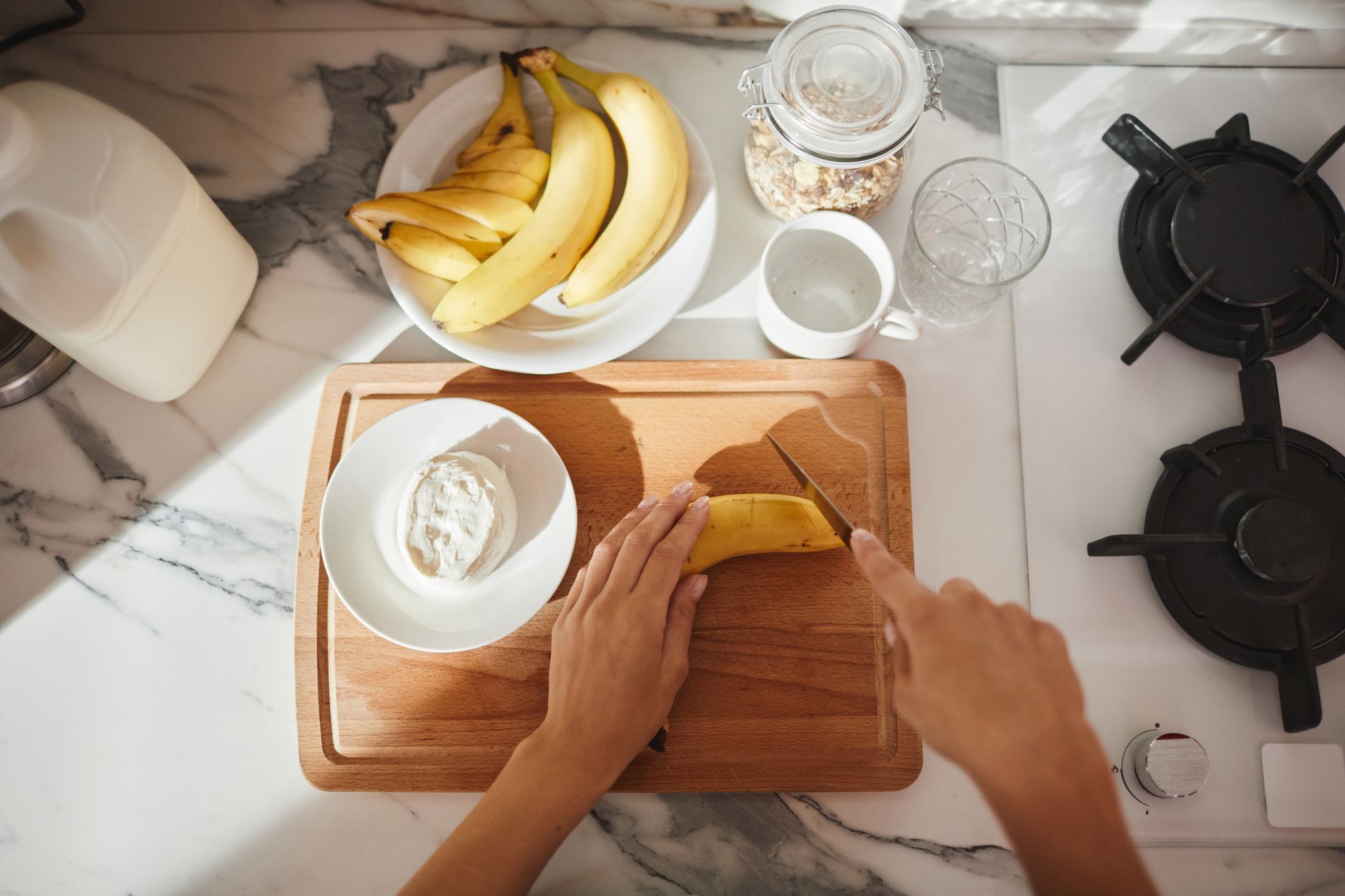 Manos cortando rodajas de plátano junto a un frasco de avena, un bol de yogur y leche sobre la mesada de la cocina