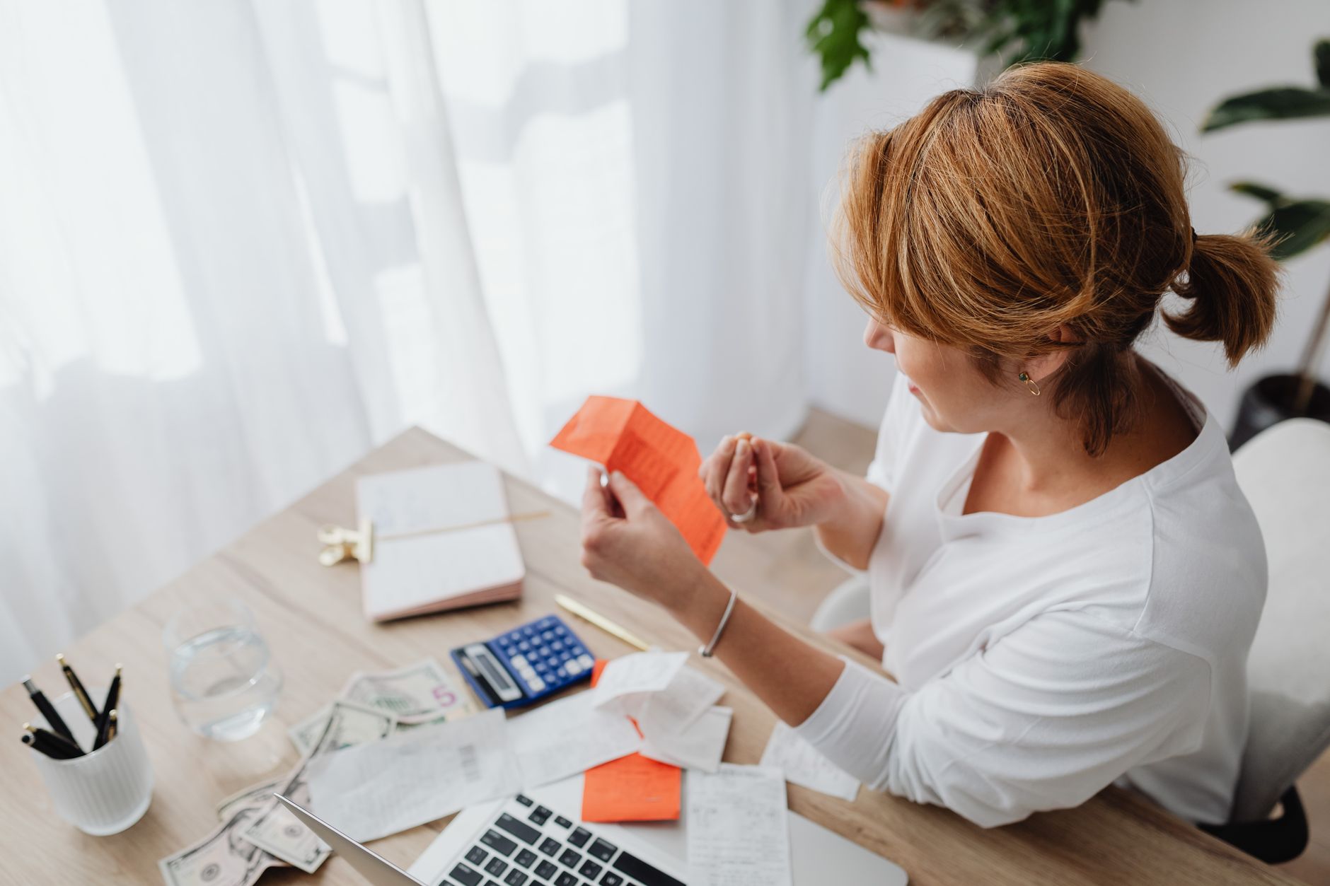 Mujer revisando recibos y facturas con una calculadora, billetes y una laptop en la mesa de su casa