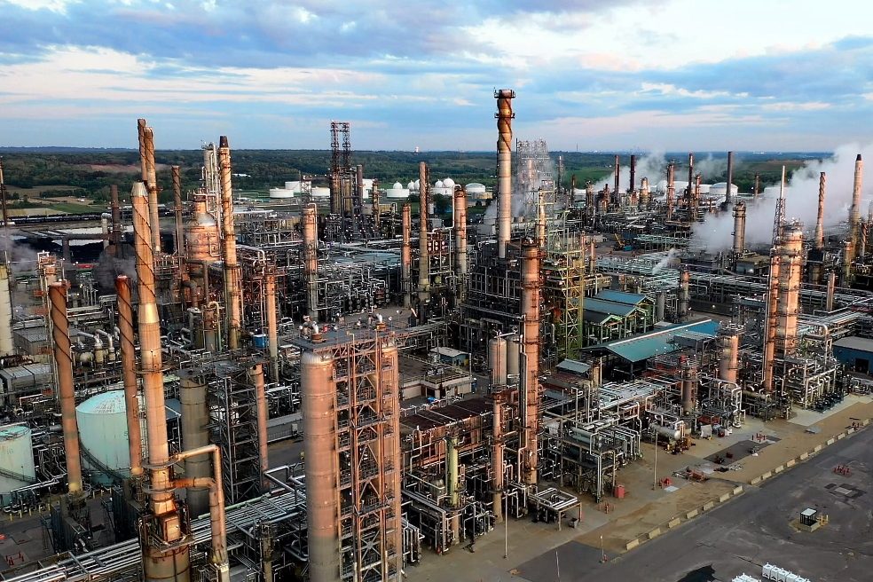 Vista aérea de una gran refinería de petróleo con numerosas torres y tuberías emitiendo vapor bajo un cielo nublado