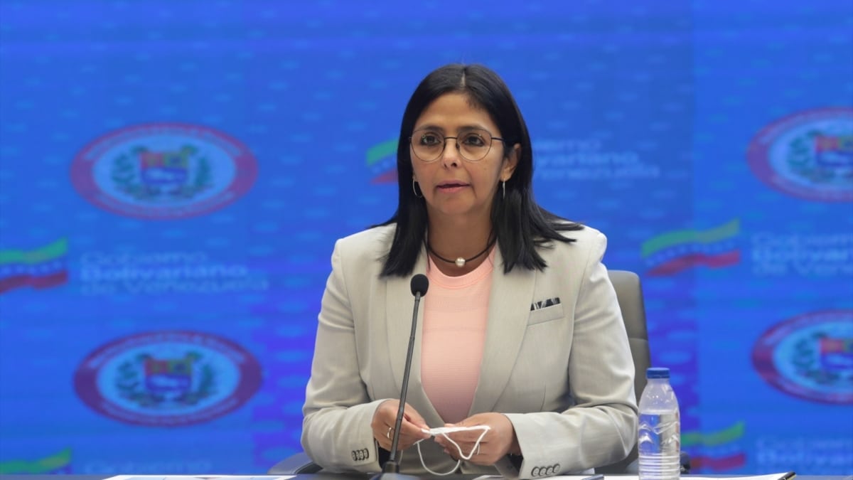 Mujer con gafas, chaqueta clara y blusa rosa habla en un atril con micrófono frente a un fondo azul con el escudo de Venezuela