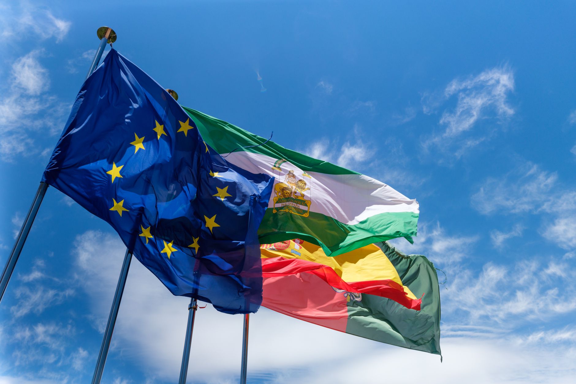 Banderas de la Unión Europea, de España y de Andalucía ondeando juntas en sus mástiles bajo un cielo azul