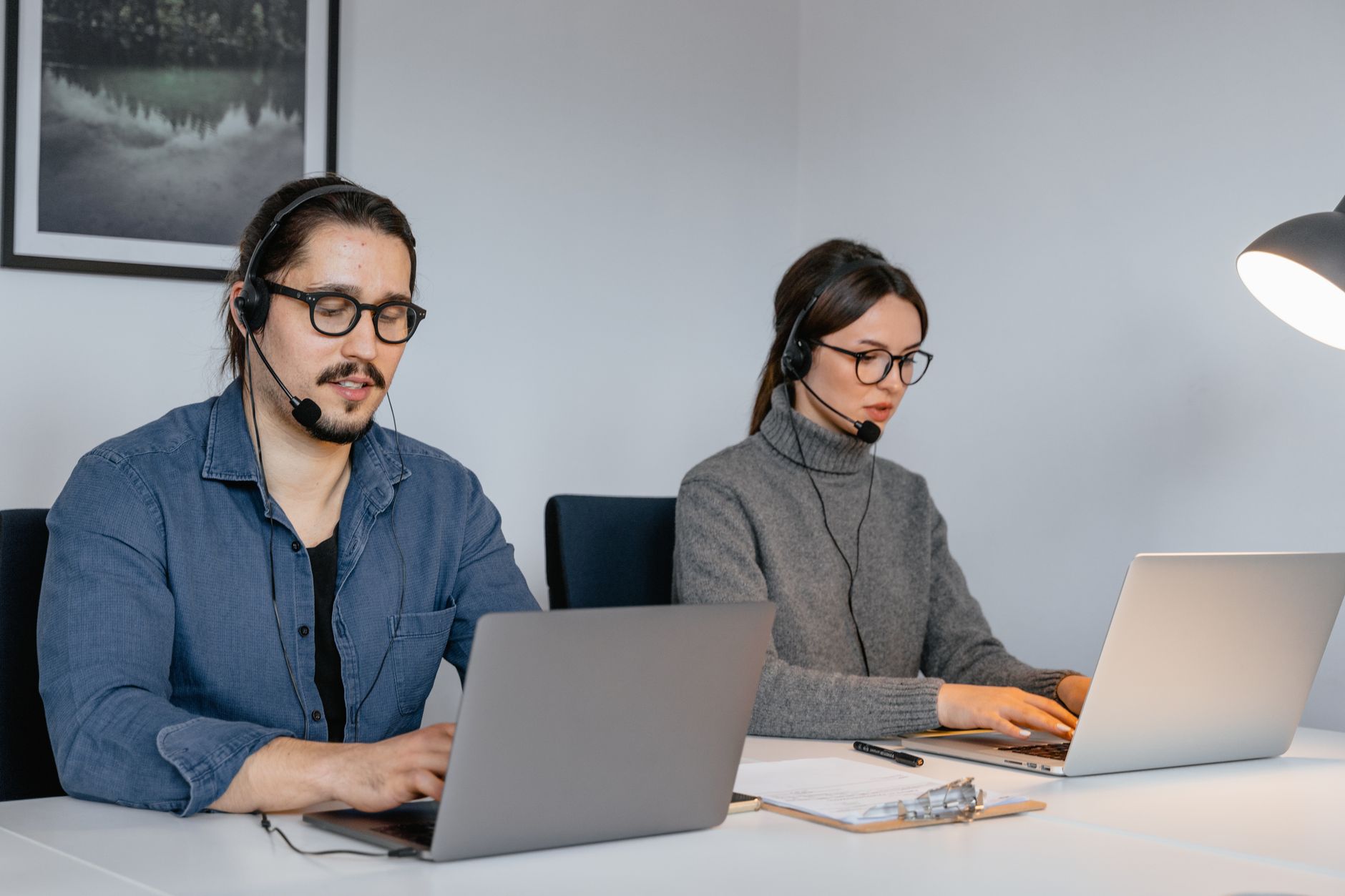 Dos empleados con auriculares de diadema trabajan concentrados en sus portátiles en una oficina de atención al cliente