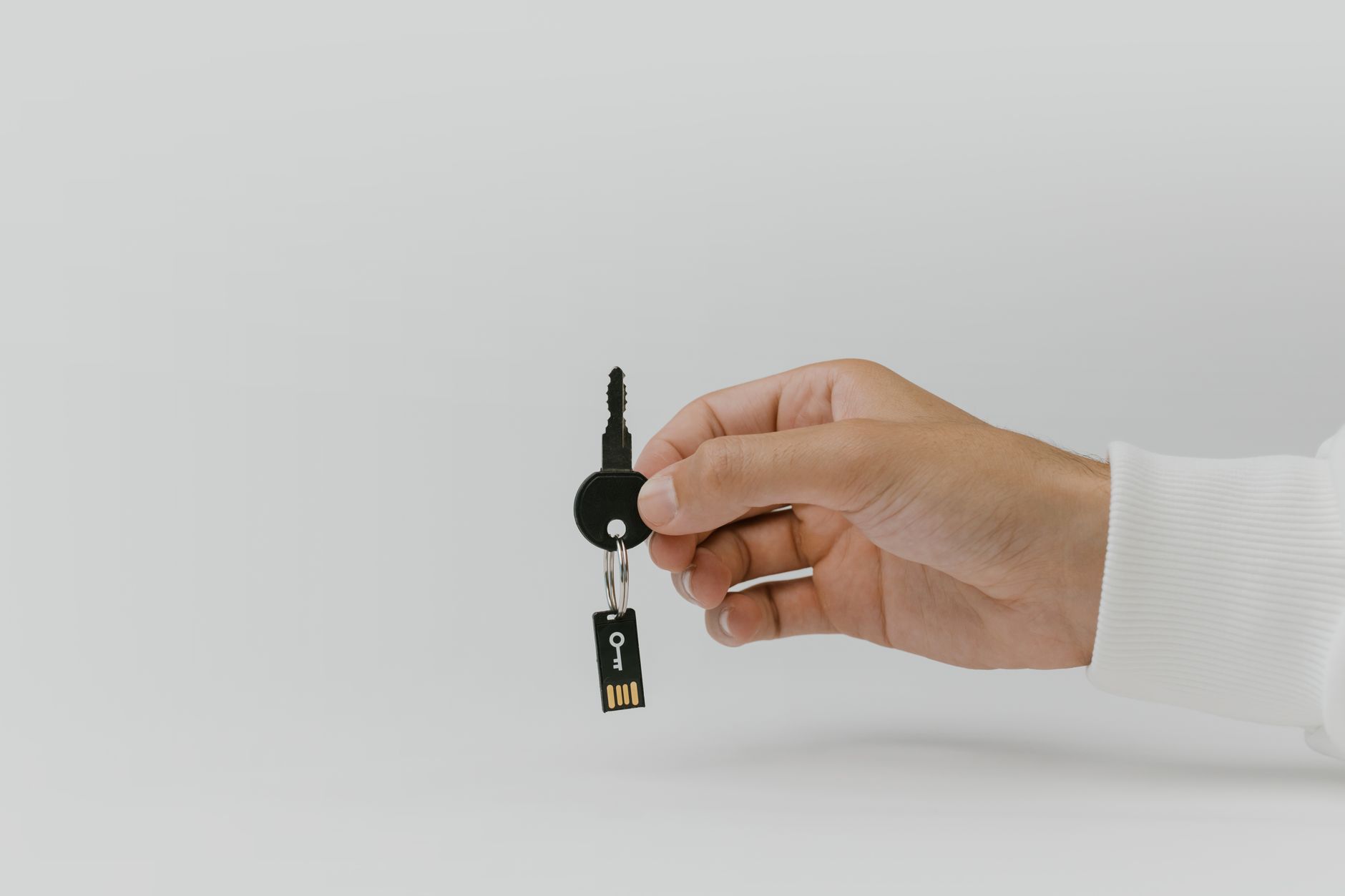 Una mano sostiene un llavero con una llave negra y una pequeña memoria USB de seguridad sobre un fondo gris claro