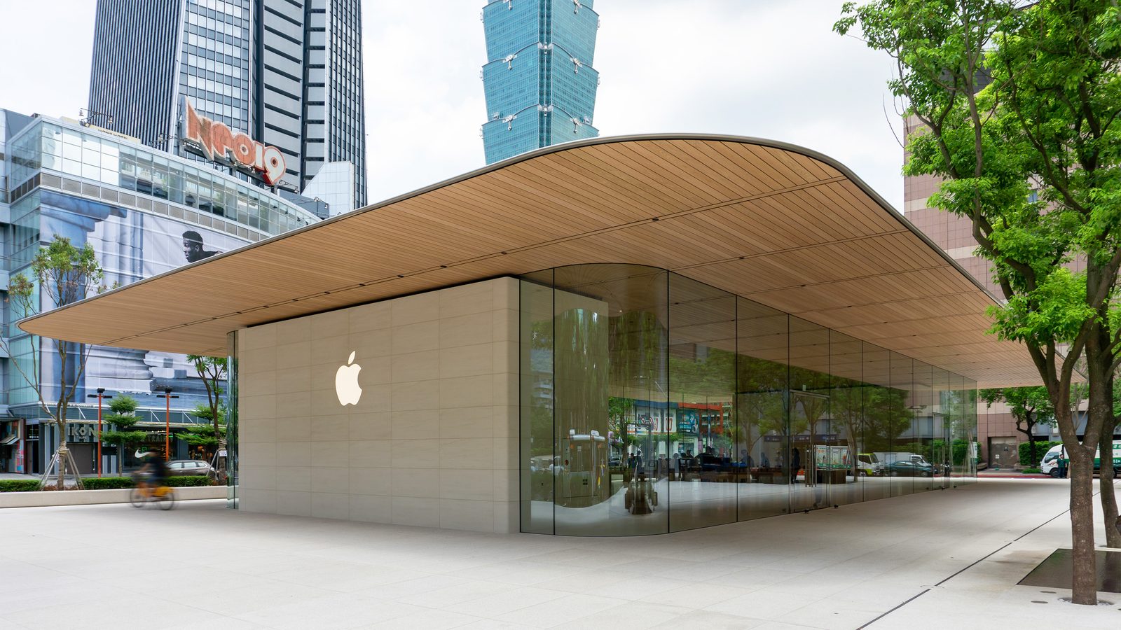 Tienda moderna de Apple con techo de madera y paredes de cristal en una plaza urbana rodeada de rascacielos