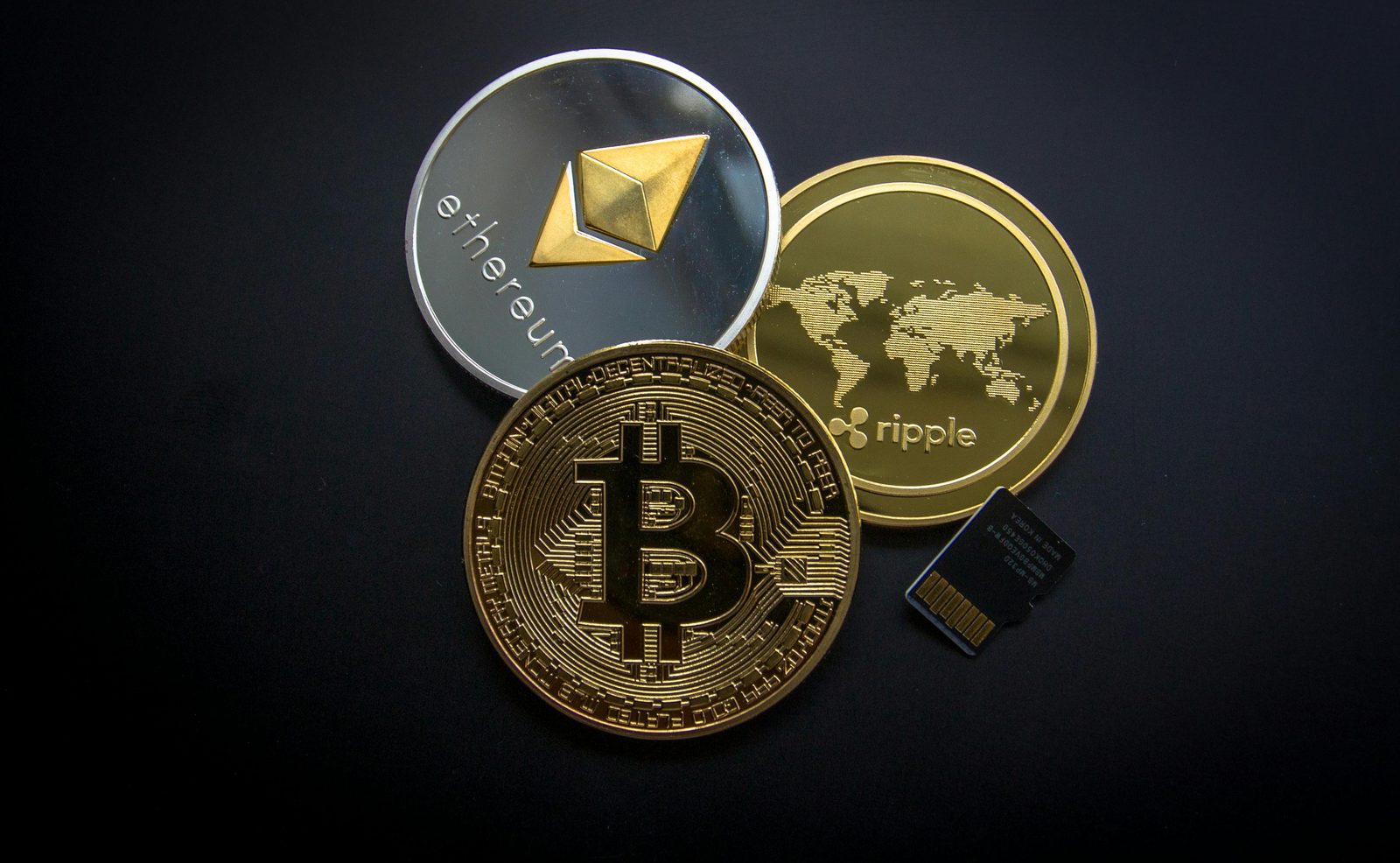 Monedas físicas de Bitcoin, Ethereum y Ripple junto a una tarjeta micro SD sobre una superficie oscura