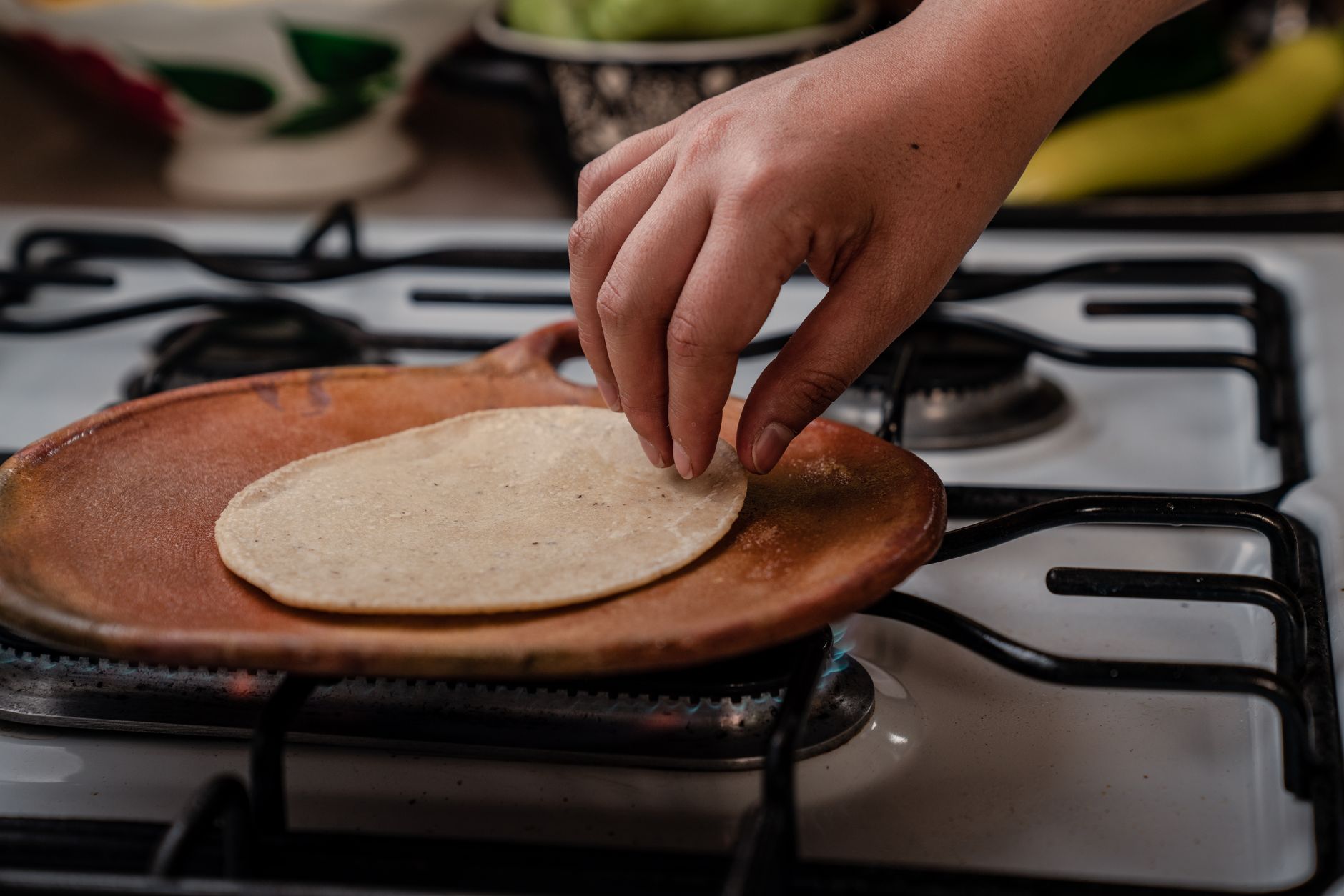 Una mano coloca una tortilla de maíz sobre un comal de barro puesto en una estufa de gas