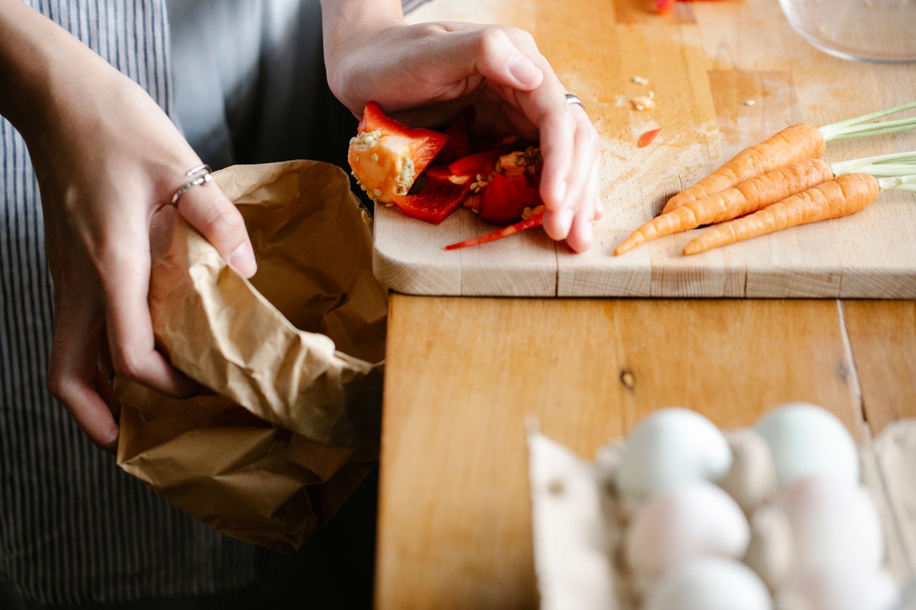 Unas manos echan a una bolsa de papel los recortes y las semillas de un pimiento, junto a zanahorias con sus tallos