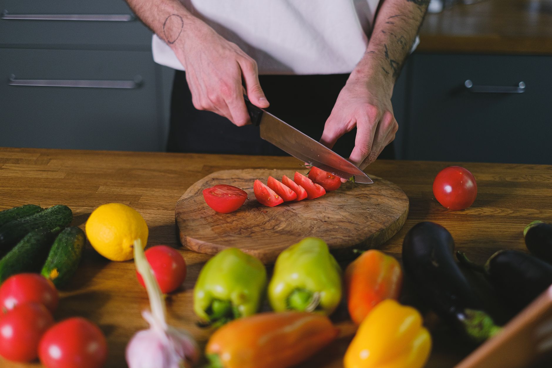 Un cocinero corta tomates en rodajas sobre una tabla de madera rodeado de pimentones, pepinos y berenjenas frescas