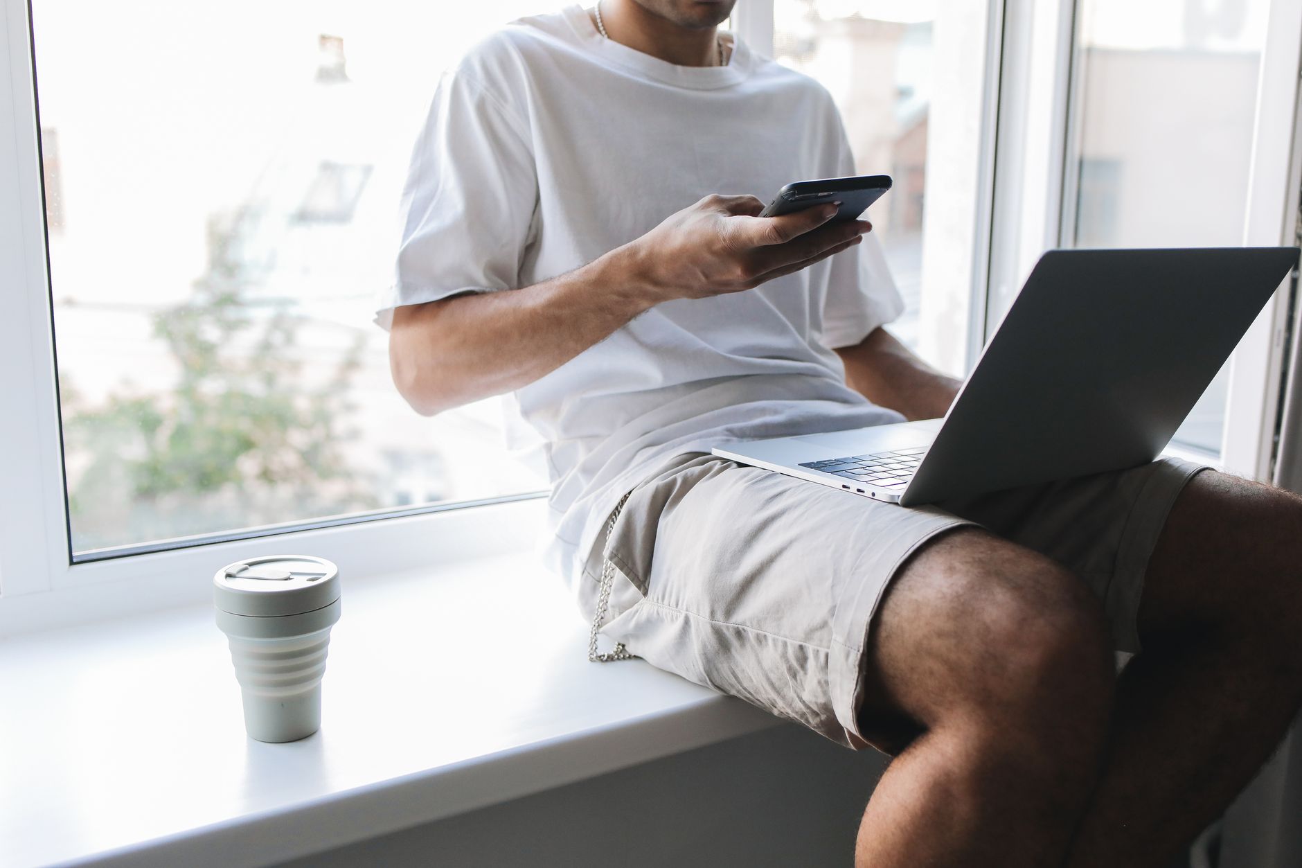 Persona usando una aplicación en el teléfono con la computadora portátil abierta sobre las piernas junto a la ventana