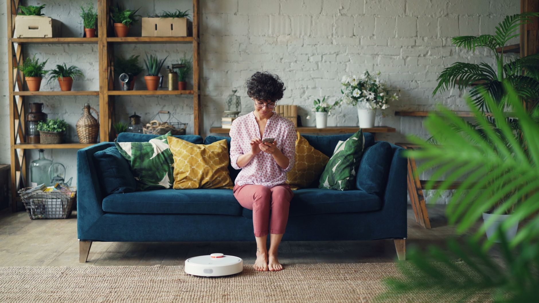 Mujer sentada en el sofá de su sala usa el teléfono mientras una aspiradora robot limpia el piso
