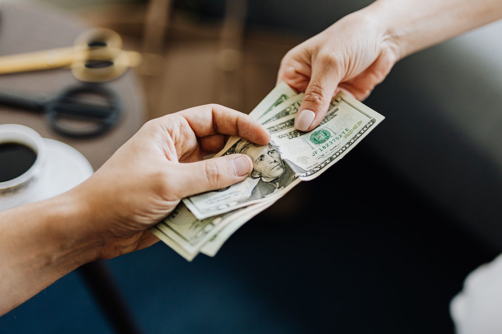 Manos entregando varios billetes de veinte dólares a otra persona sobre una mesa de madera