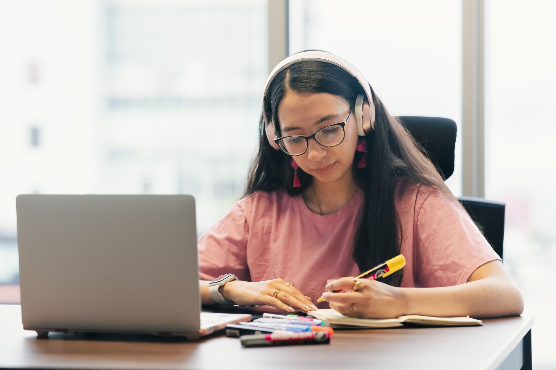 Una joven con audifonos toma apuntes en su cuaderno mientras sigue una clase en la computadora