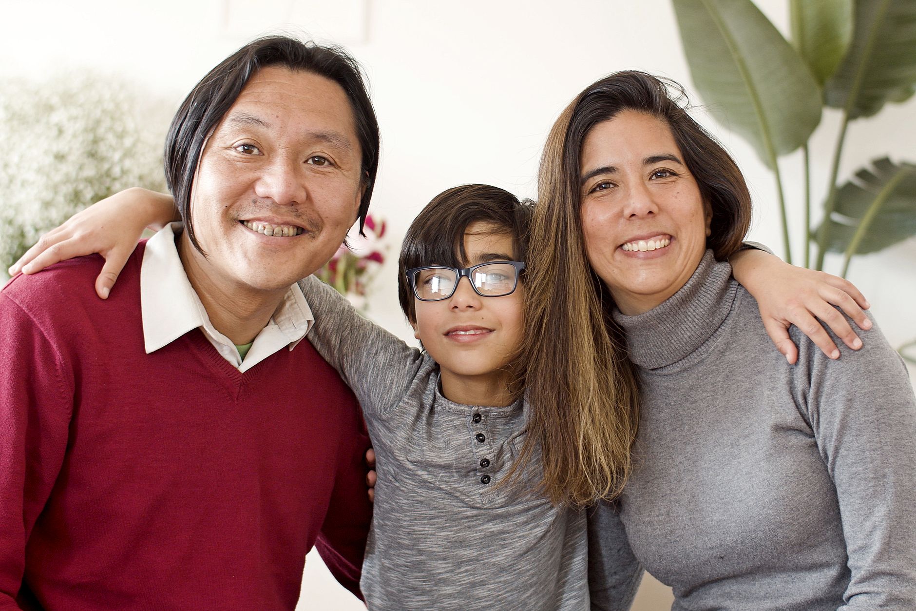 Una madre latina, un padre y su hijo con lentes posan abrazados y sonriendo en la sala de su casa