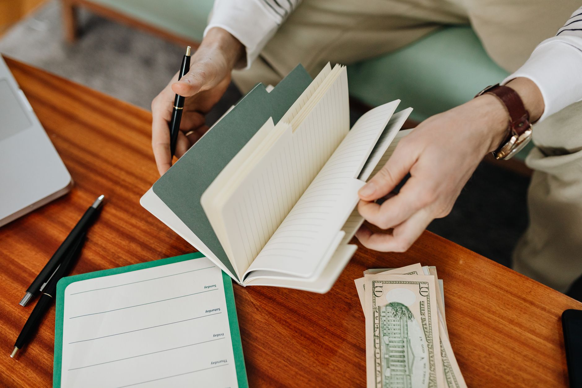 Persona repasando su cuaderno de notas con boligrafo junto a unos billetes de dolar sobre una mesa de madera
