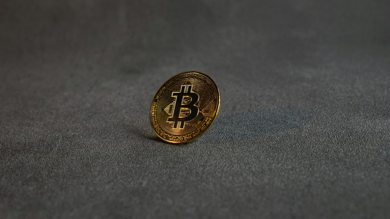 Una moneda dorada de Bitcoin con el logo B en relieve, apoyada de lado sobre una superficie gris texturizada
