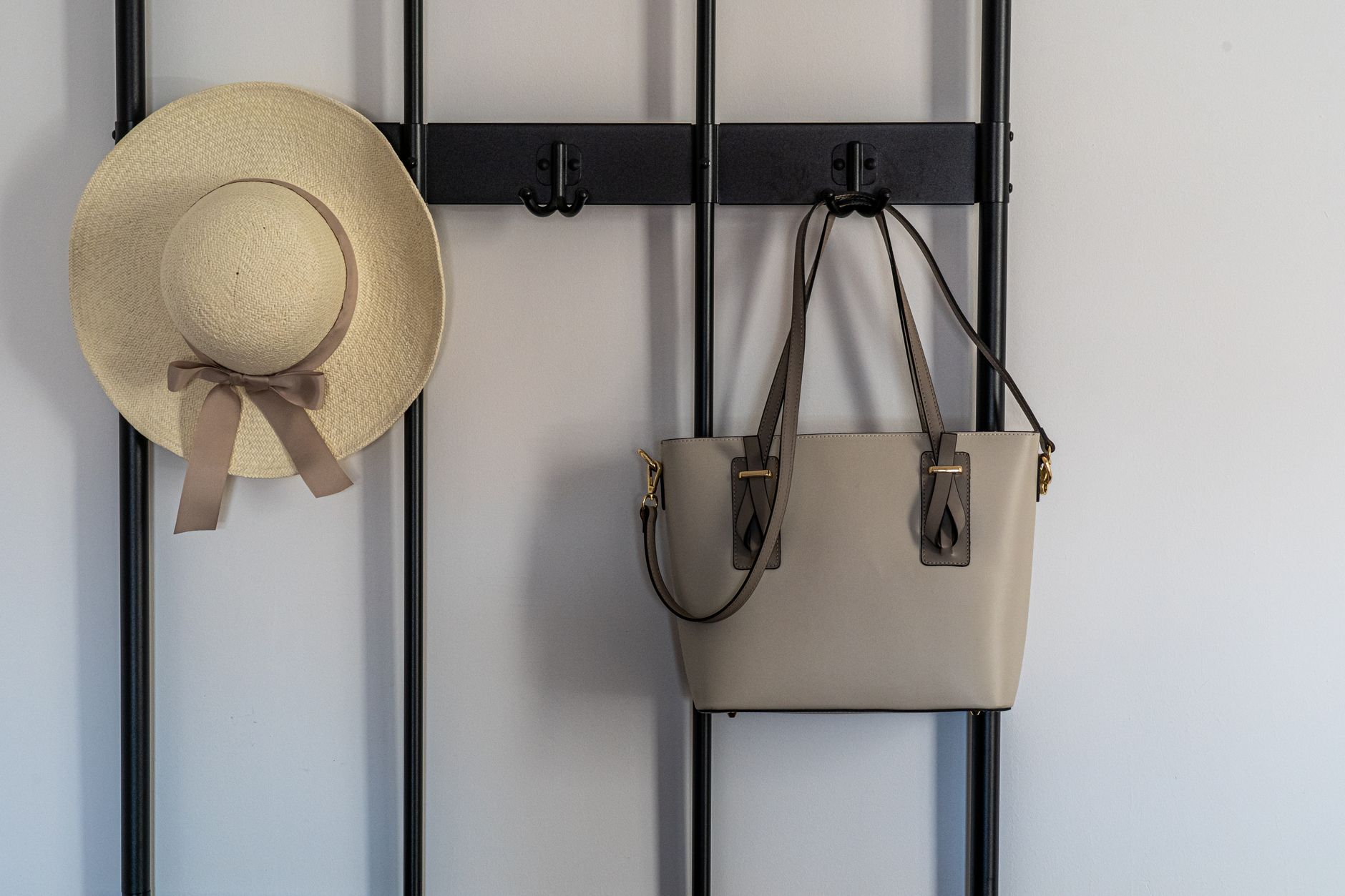 Un sombrero de paja y un bolso de mano colgados de los ganchos de un perchero de barras negras apoyado contra una pared blanca