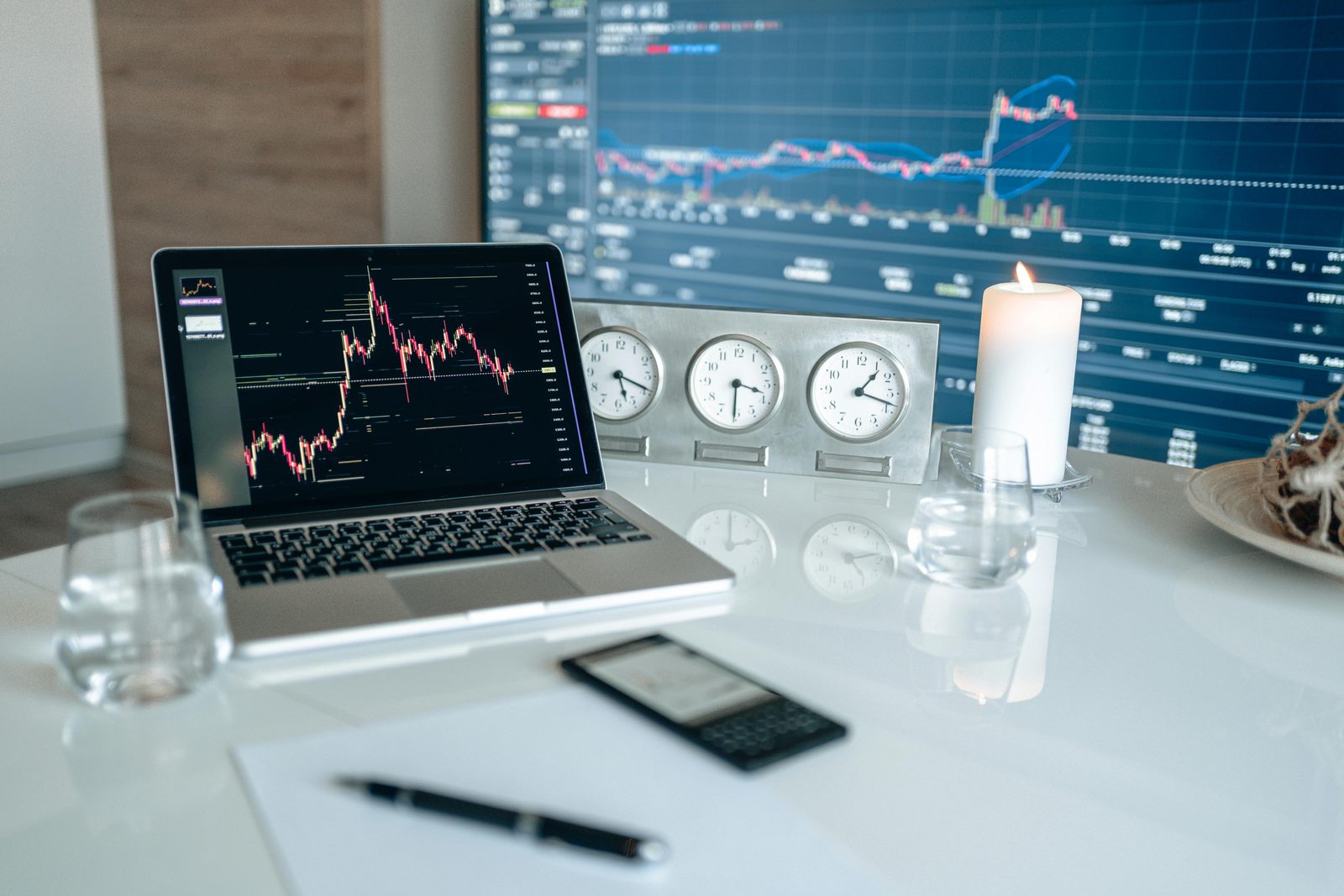 Mesa de trading con portátil, velas y tres relojes; en el monitor se ven gráficos bursátiles
