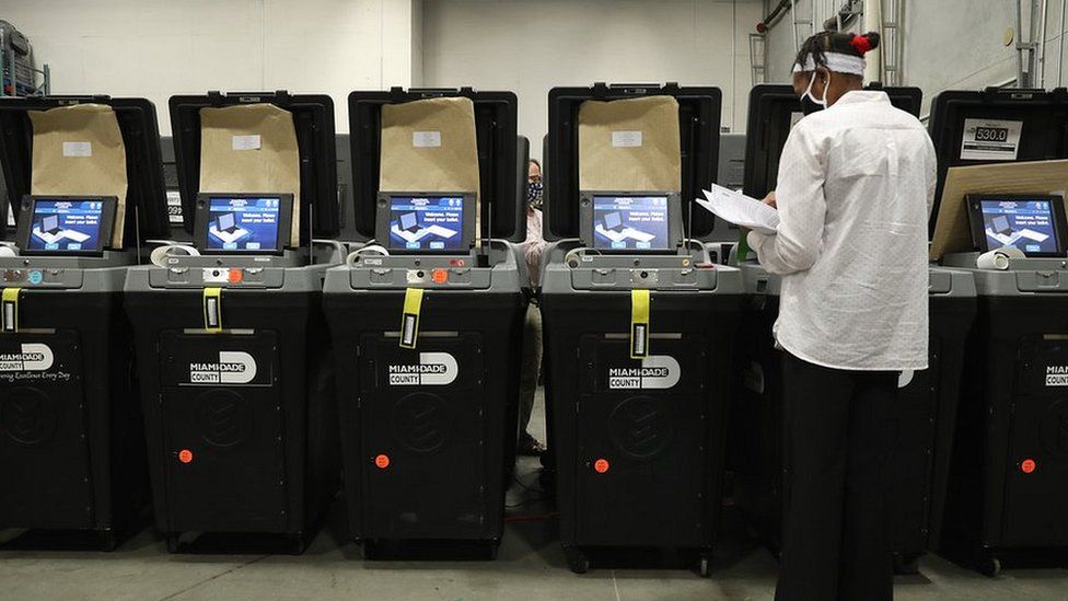 Persona revisando papeles frente a una fila de máquinas de votación con el rótulo Miami-Dade County en un centro electoral