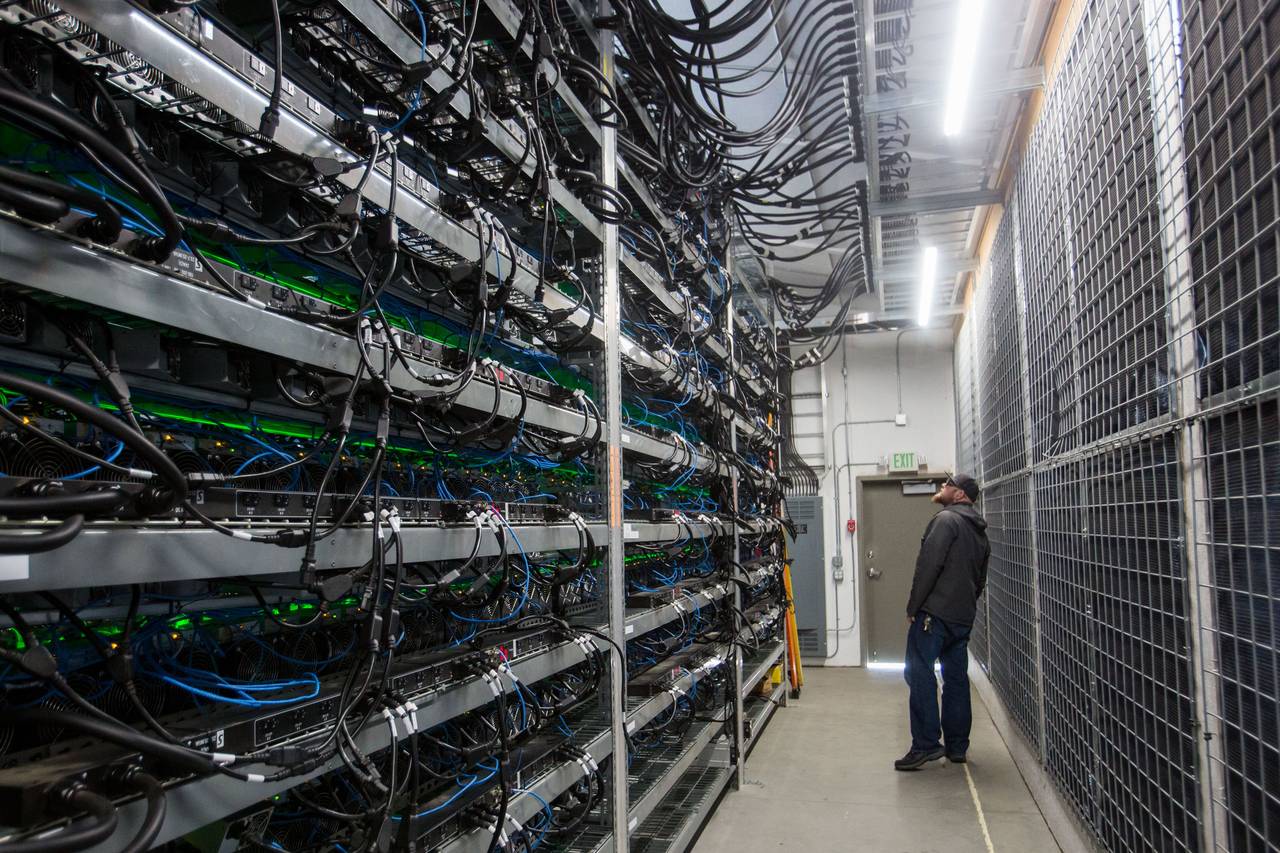 Hombre de pie en un pasillo de un centro de datos observando filas de servidores llenos de cables y un letrero de Exit
