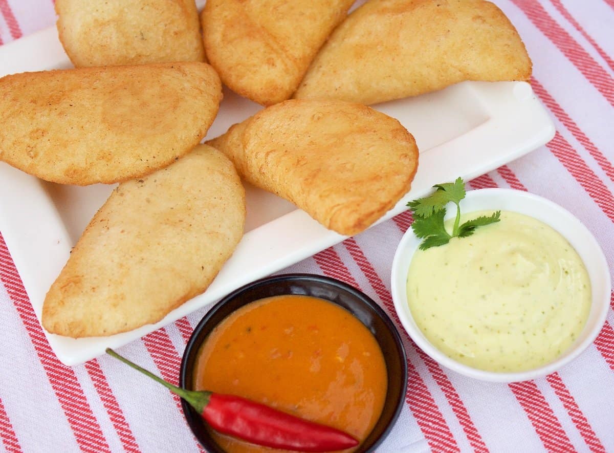 Seis empanadas fritas doradas en un plato blanco, con dos cuencos de salsa y un chile rojo fresco sobre un mantel a rayas