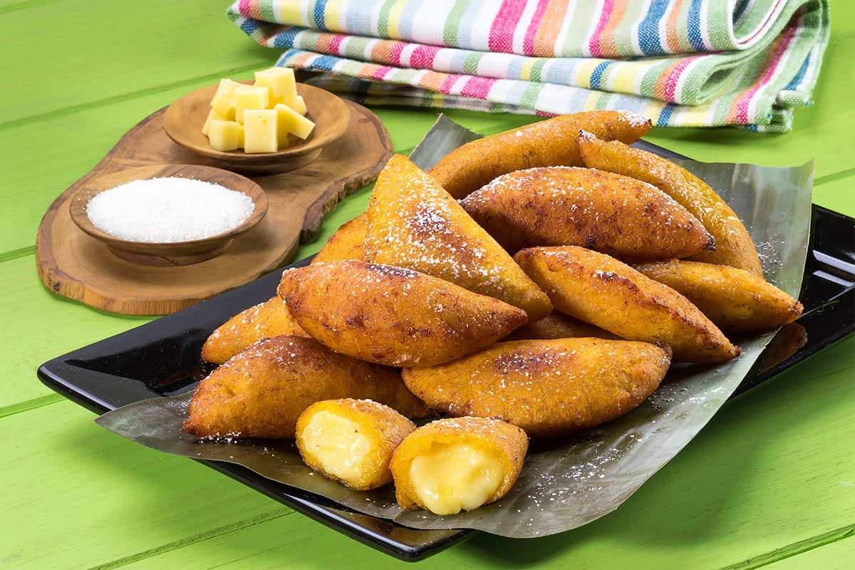Empanadas fritas de plátano y queso espolvoreadas con azúcar sobre hojas en un plato negro, con queso en cubos al lado