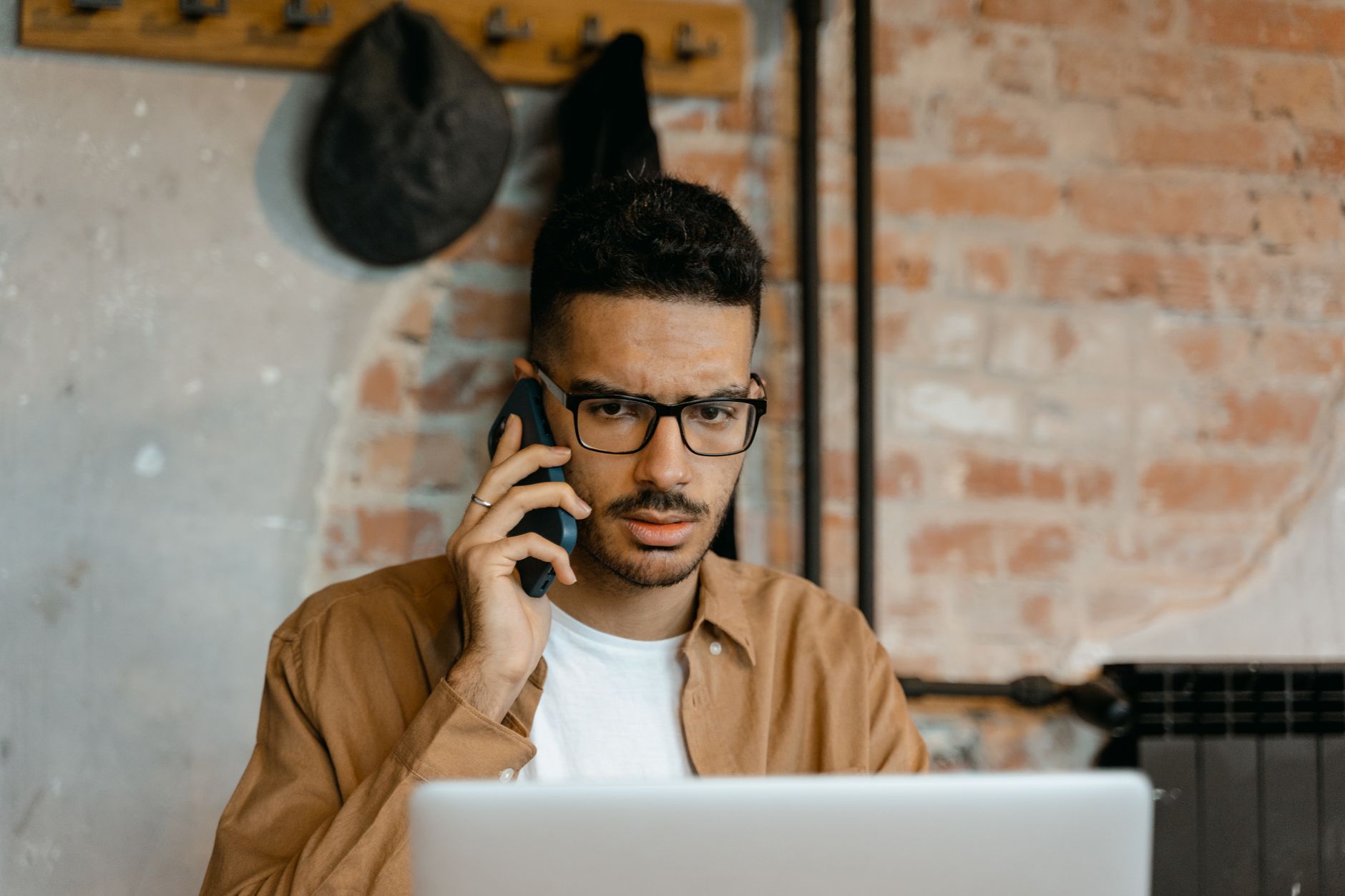 Hombre con gafas escucha con gesto serio una llamada telefónica frente a su portátil, haciendo preguntas antes de comprar