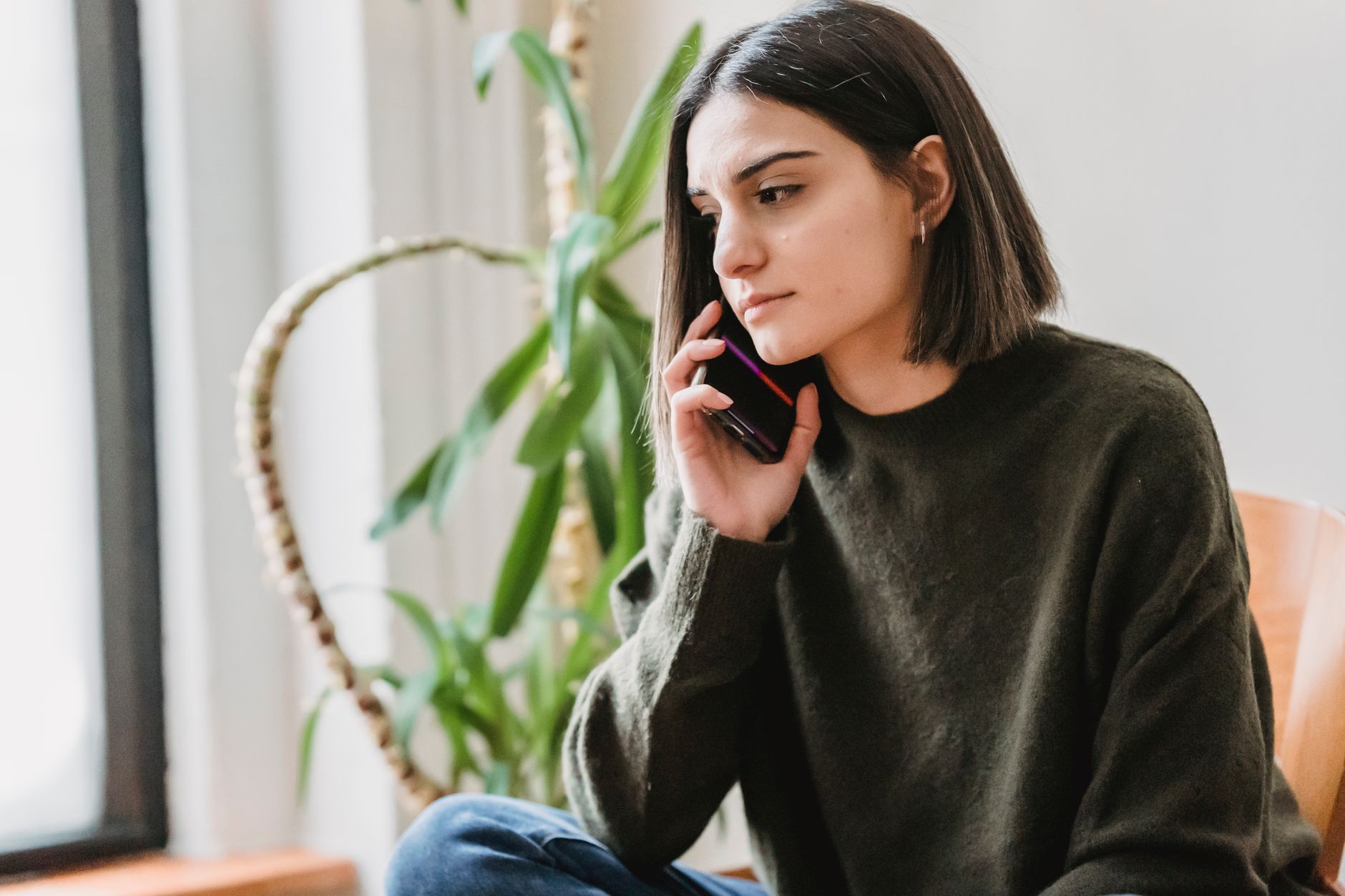 Mujer joven sentada en un sillón de su casa hablando por teléfono con gesto serio