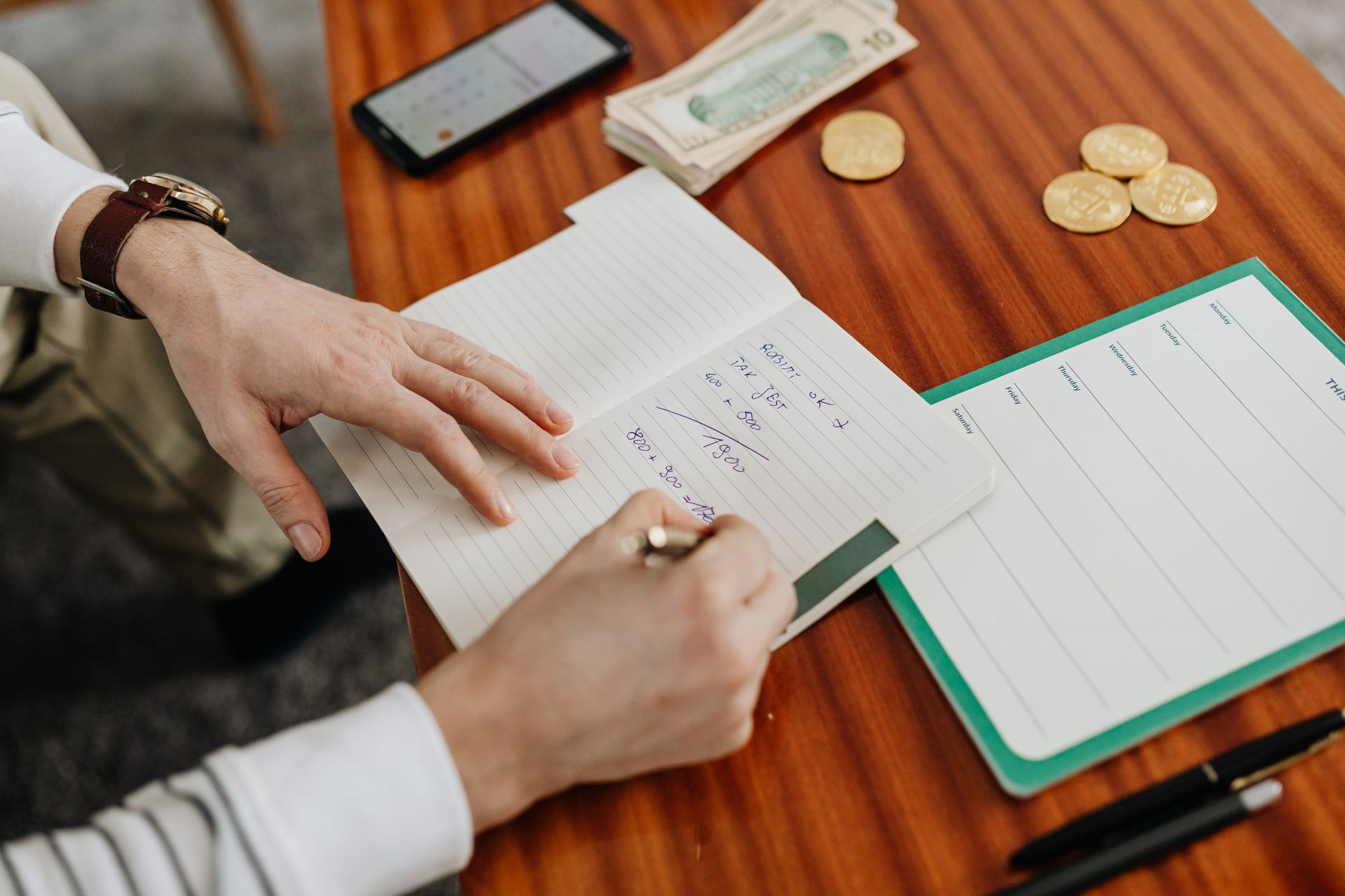 Manos anotan cuentas a mano en una libreta junto a billetes, monedas y una planilla en blanco