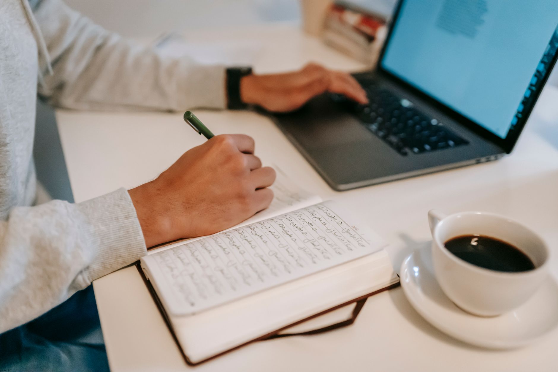 Persona tomando apuntes a mano en una libreta mientras teclea en una laptop, con una taza de café al lado