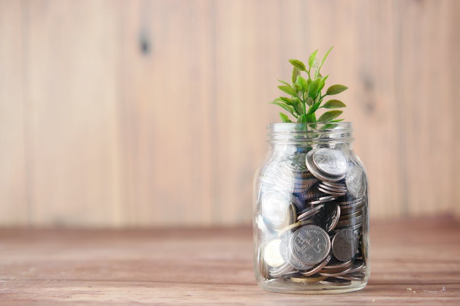 Tarro de cristal lleno de monedas con una pequeña planta verde brotando de ellas, colocado sobre una mesa de madera clara