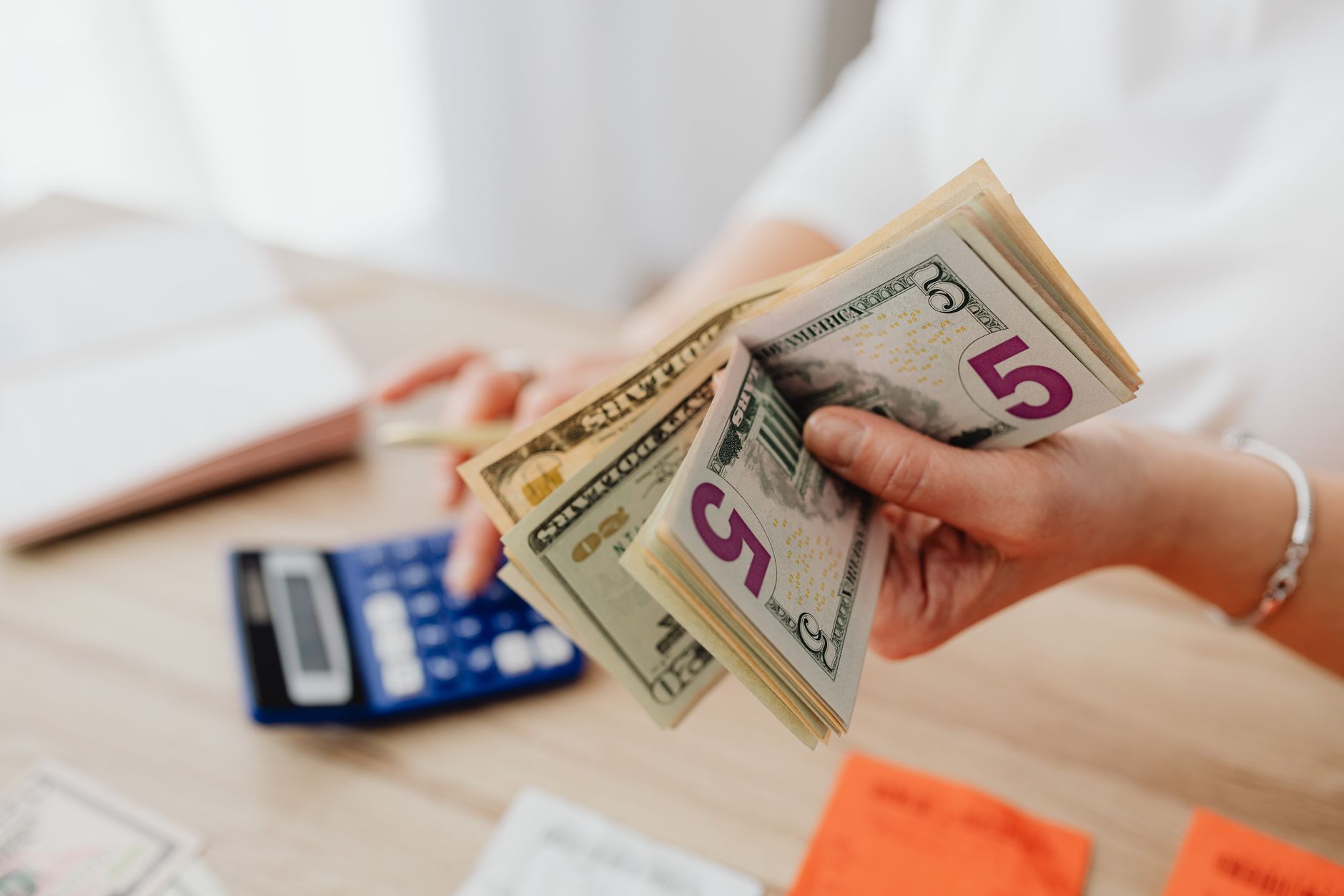 Manos contando billetes de dolar junto a una calculadora azul sobre un escritorio de madera clara