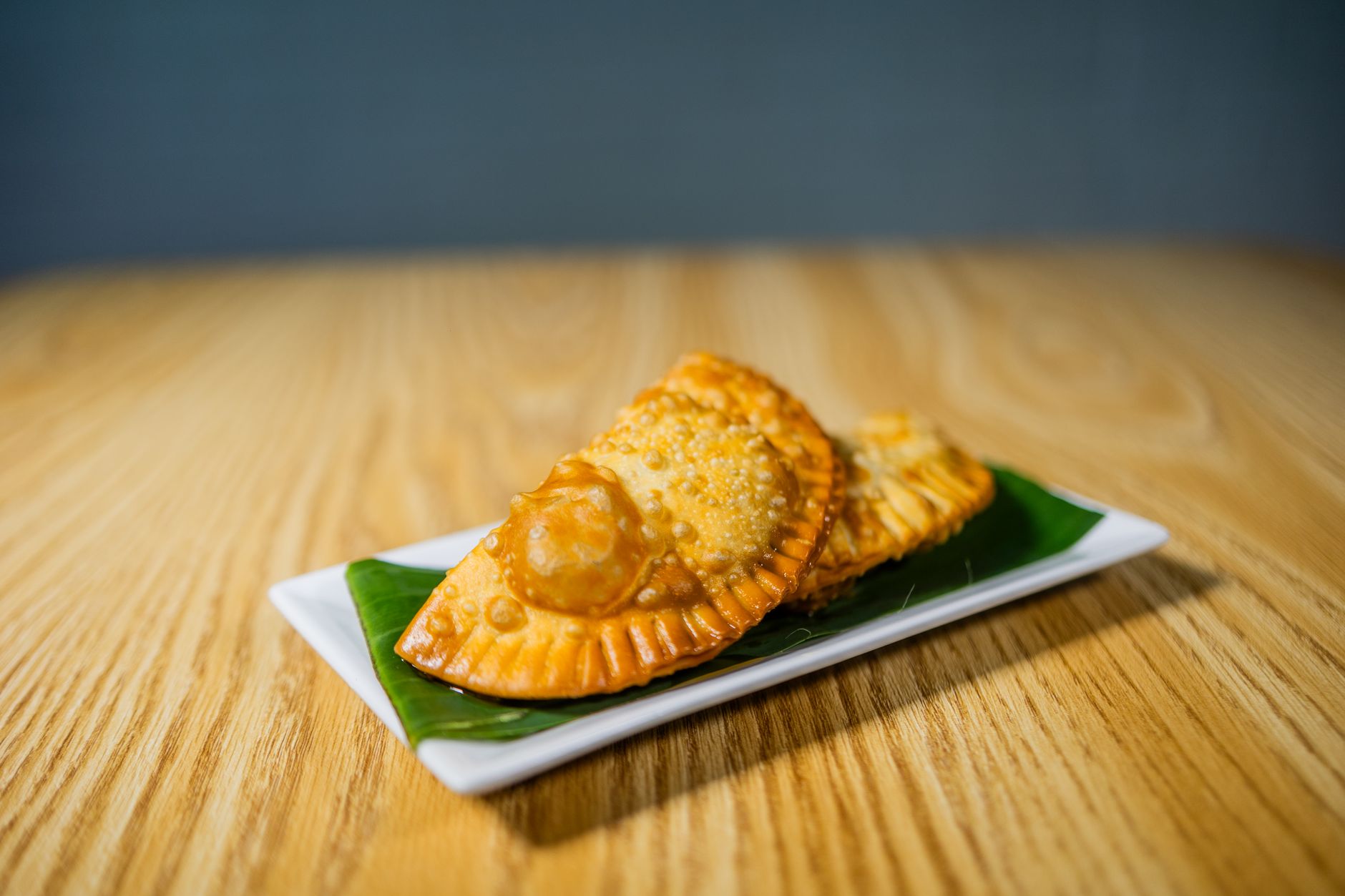 Dos empanadas fritas doradas sobre un plato blanco forrado con una hoja de plátano, encima de una mesa de madera clara