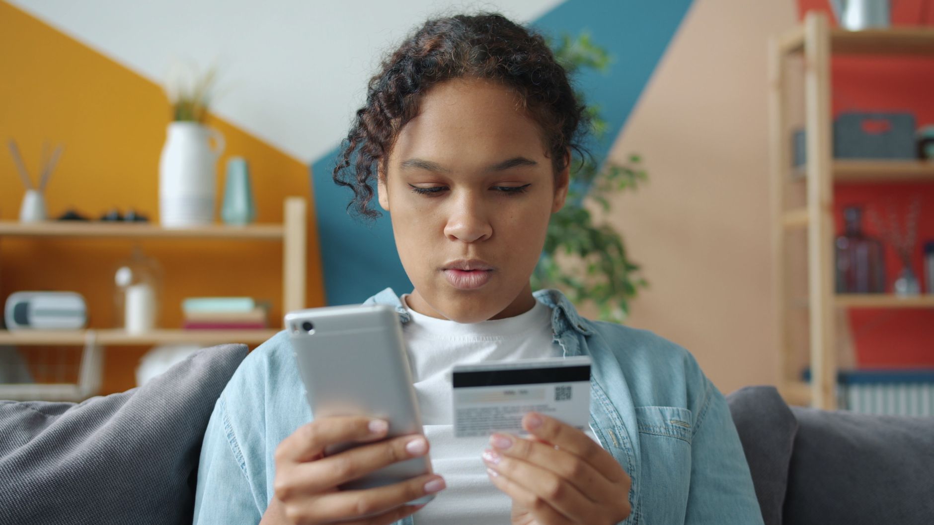 Una joven revisa la app de envío de dinero en el móvil con la tarjeta de su cuenta bancaria en la mano