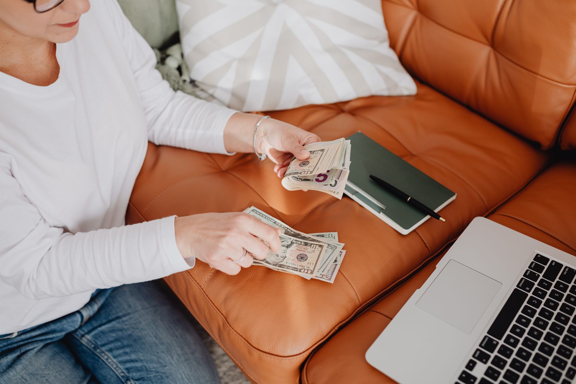 Una mujer sentada en un sofá cuenta billetes de dólar con una libreta cerrada y una laptop al lado