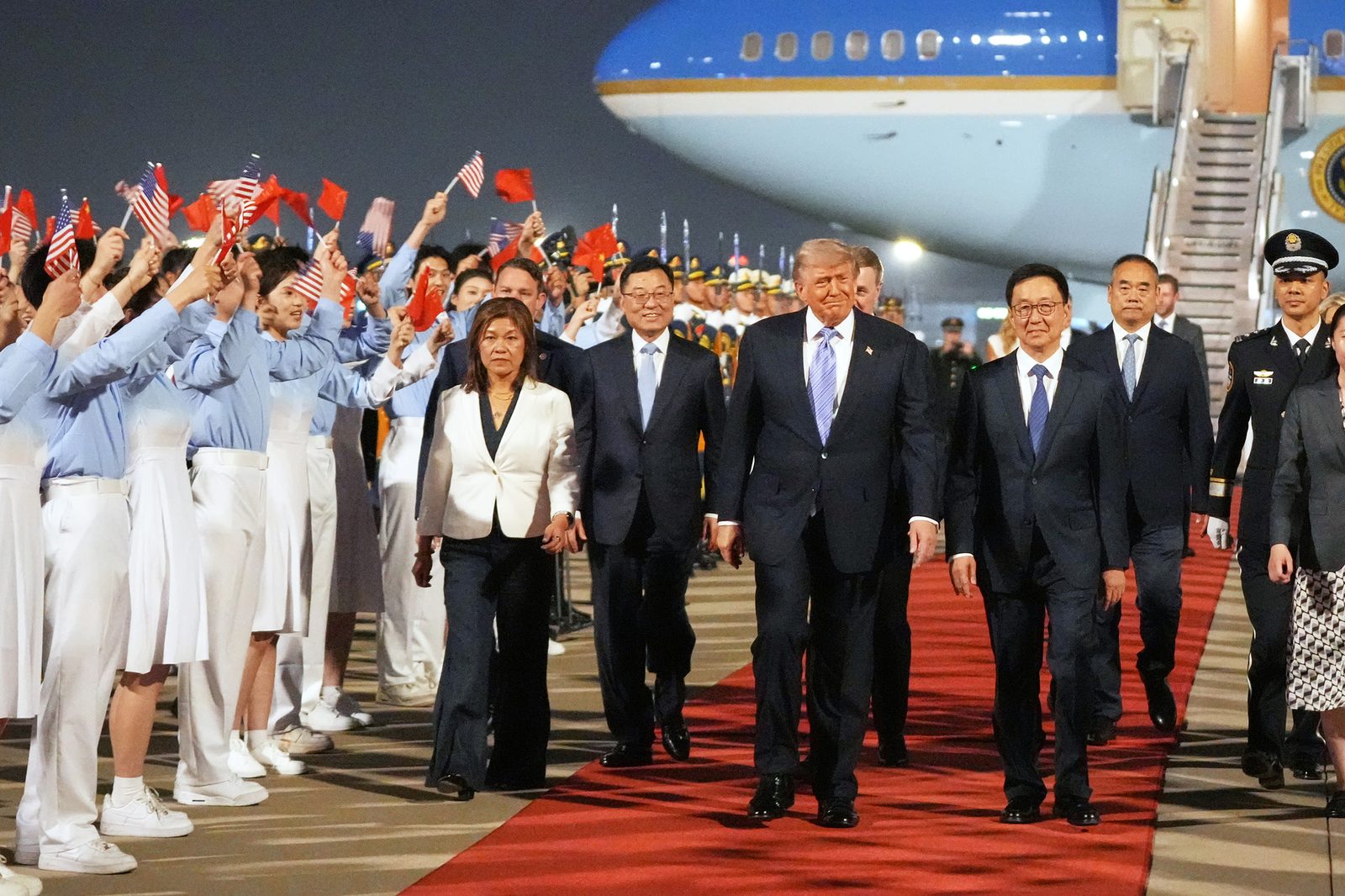 Trump y su comitiva caminan por una alfombra roja, saludados por personas con banderas de EE. UU. y China junto a un avión azul