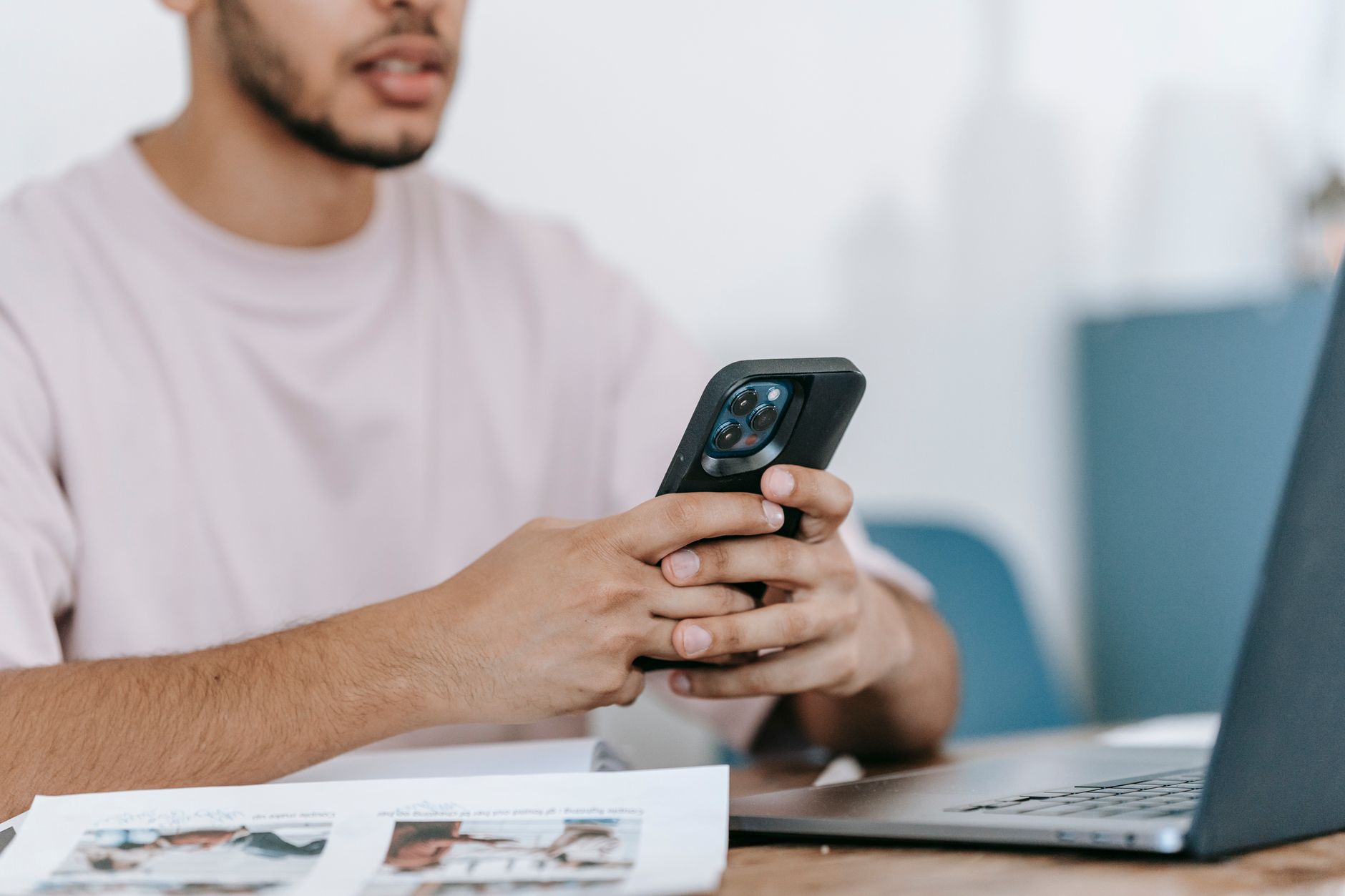 Joven en camiseta consulta el mercado en su teléfono con el portátil abierto sobre la mesa
