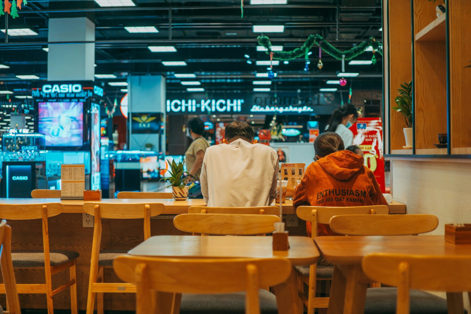 Dos personas sentadas de espaldas en un restaurante moderno con mesas de madera y letreros luminosos de fondo