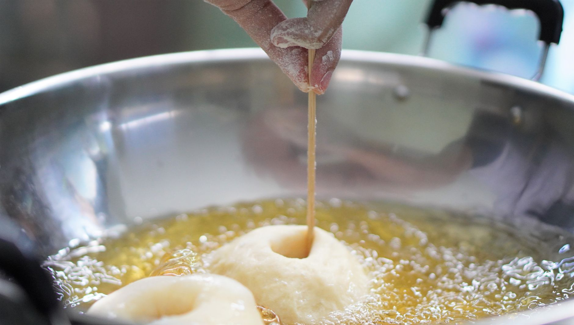 Rosquillas de masa friéndose en abundante aceite caliente, una de ellas sostenida con un palito