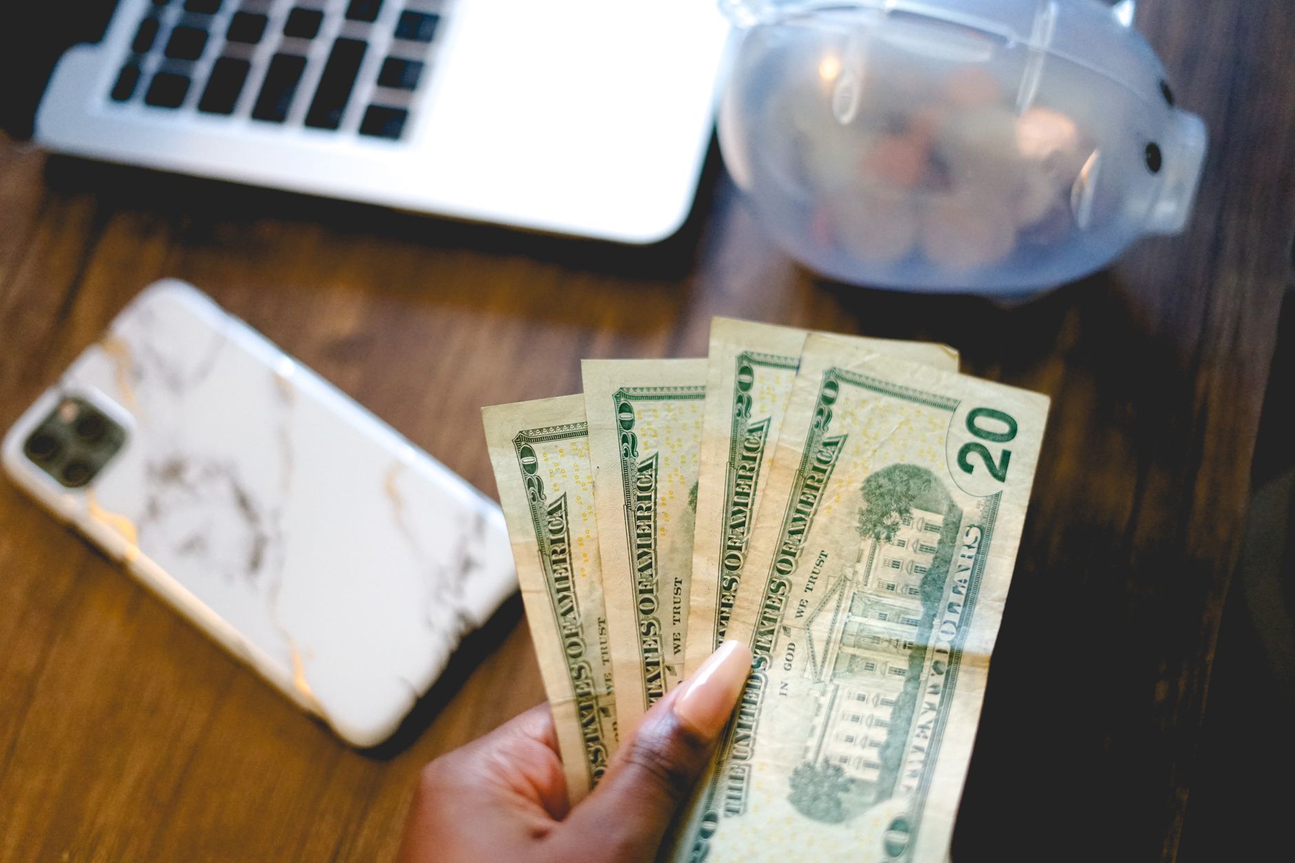 Una mano sostiene varios billetes de veinte dólares sobre una mesa de madera, junto a una laptop, un celular y una alcancía transparente con monedas
