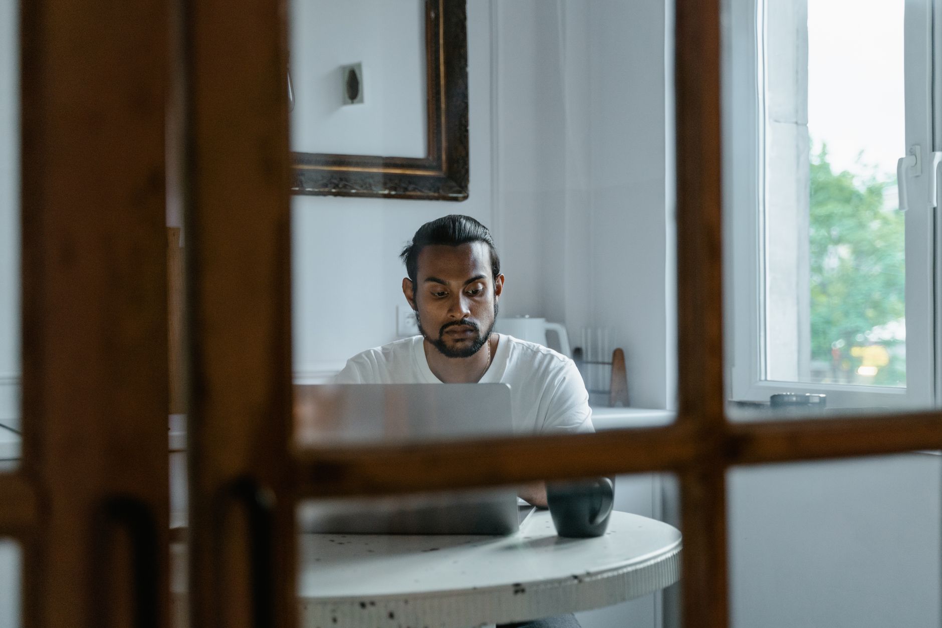 Un hombre revisa la pantalla de su portátil sentado a la mesa de su casa, junto a la ventana