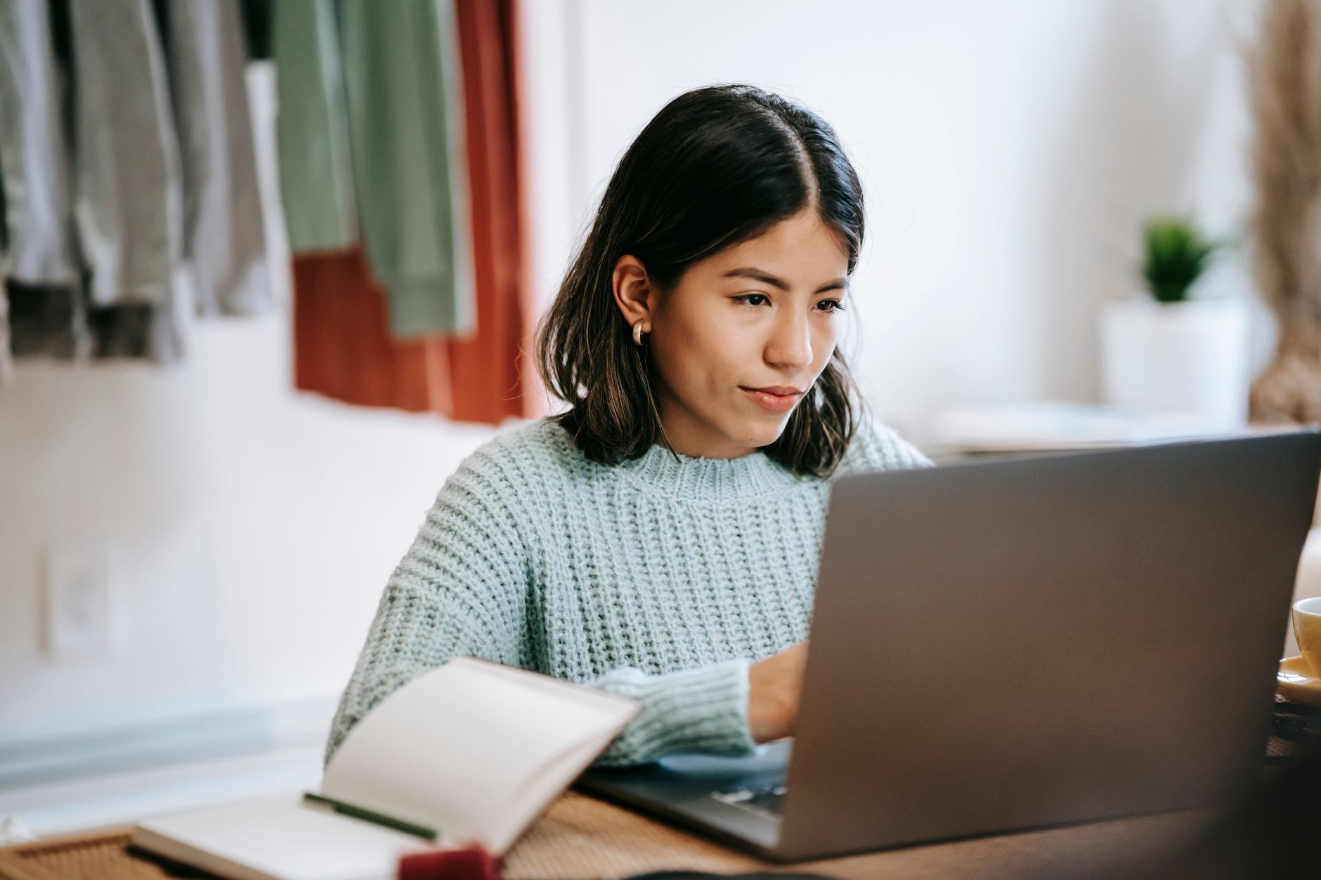 Mujer joven revisa información en su computadora portátil con un cuaderno abierto sobre la mesa