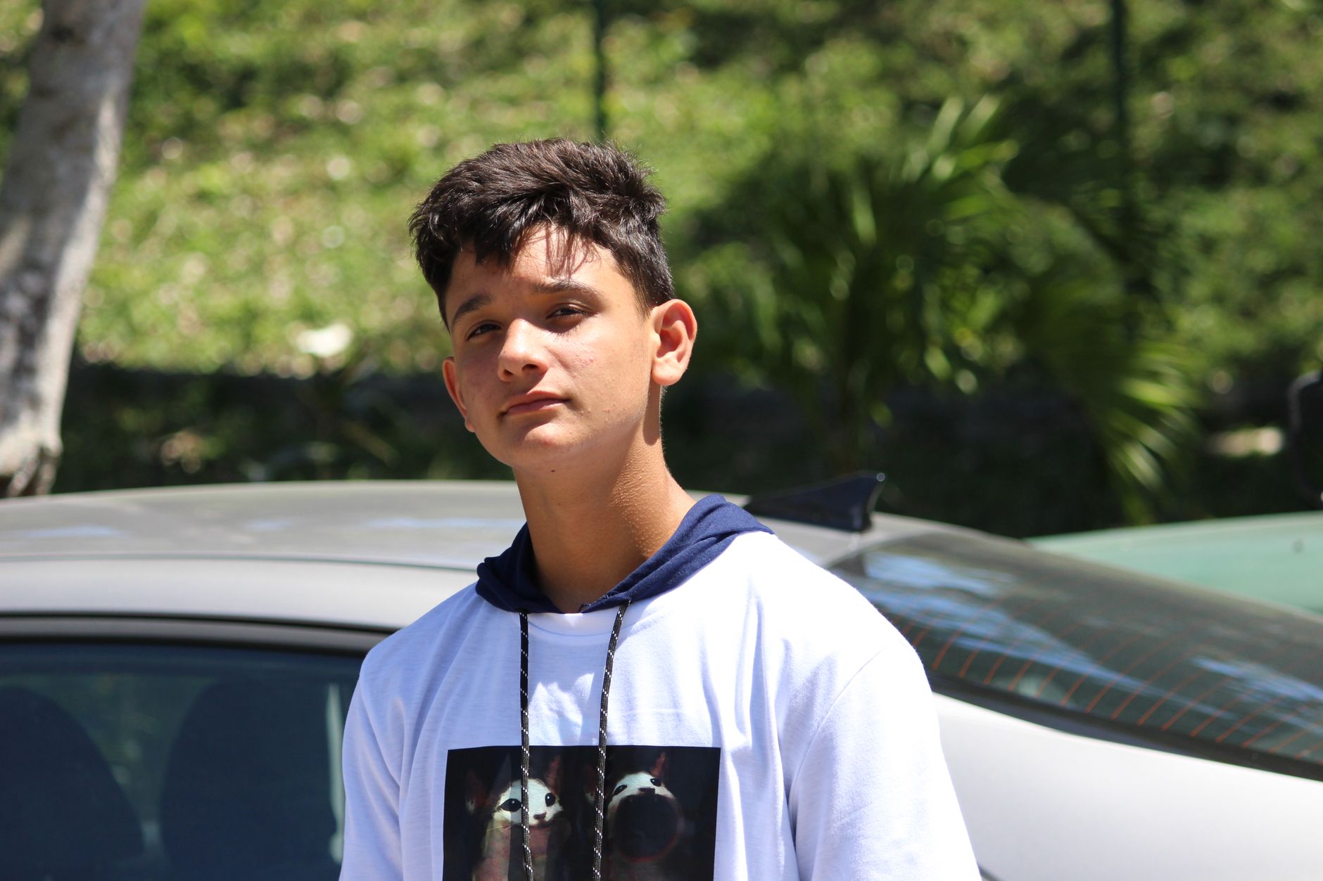 Adolescente latino de pie al sol junto a unos carros estacionados en un vecindario