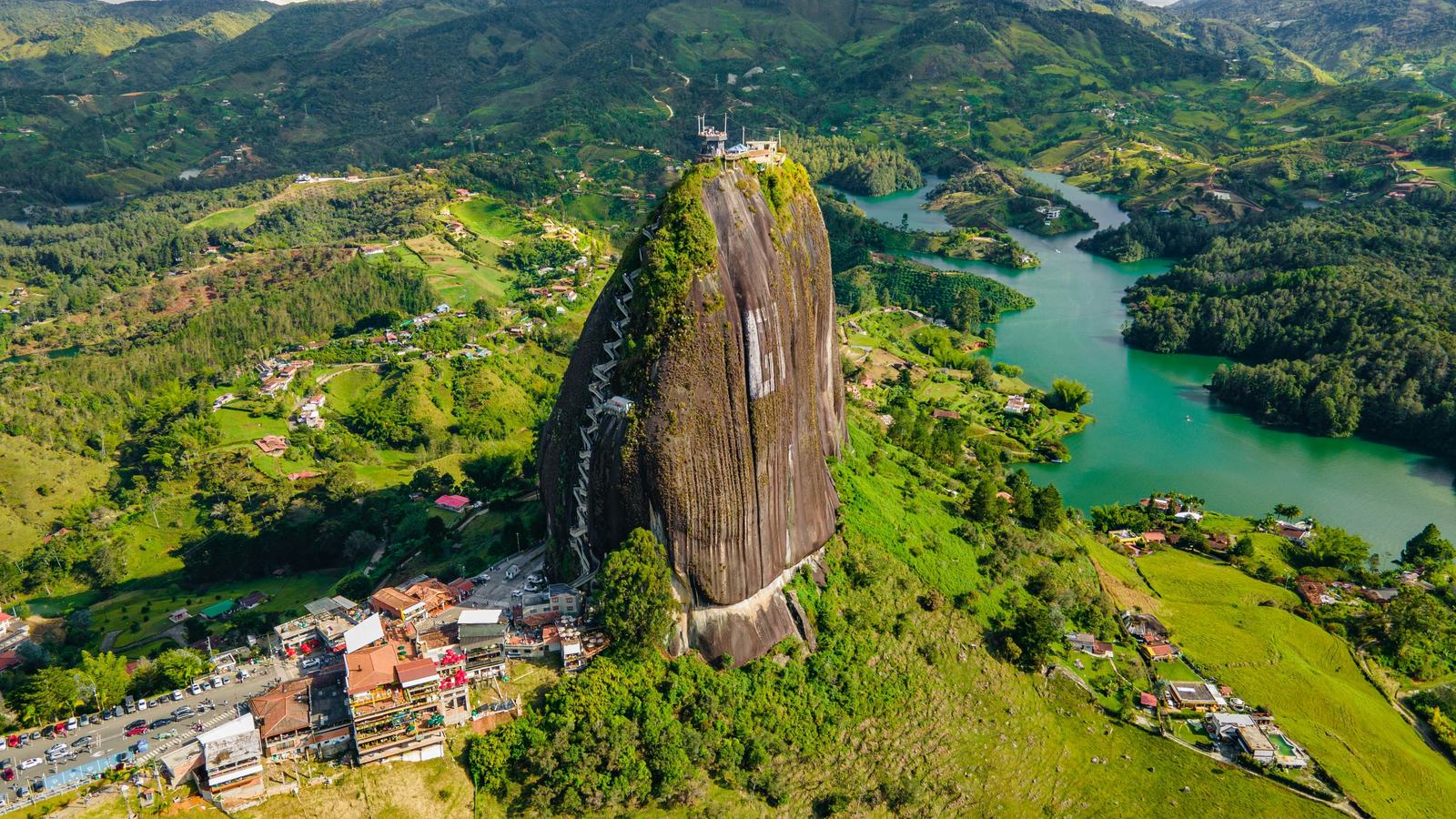 Vista aérea de la imponente Piedra del Peñol con una escalera lateral, rodeada de montañas verdes y un brillante embalse azul
