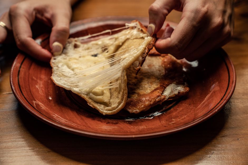 Dos manos separan una masa horneada rellena de la que cuelgan hilos finos de queso fundido sobre un plato de barro