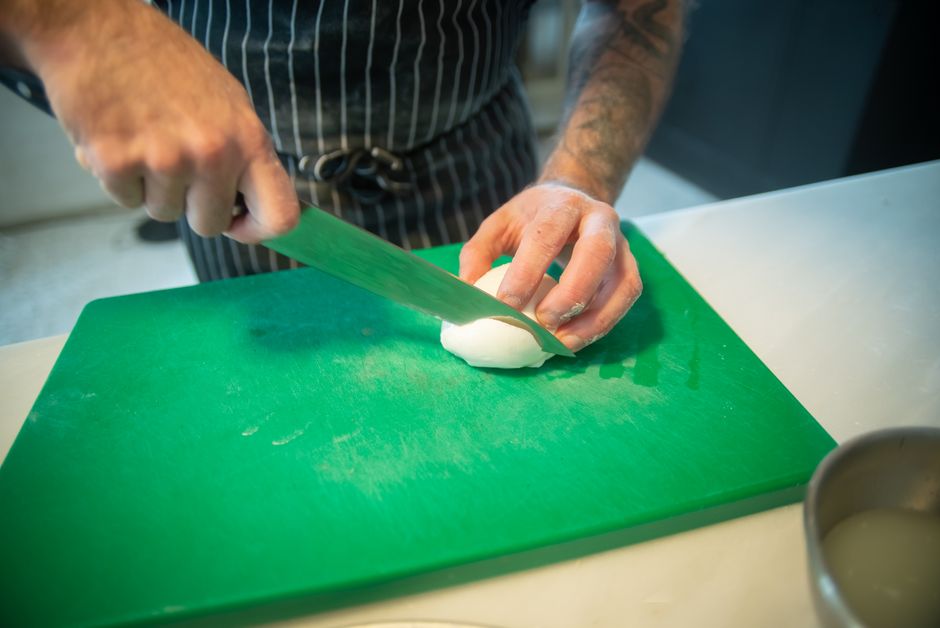 Un cocinero con delantal corta con un cuchillo una bola pequeña de queso blanco sobre una tabla verde