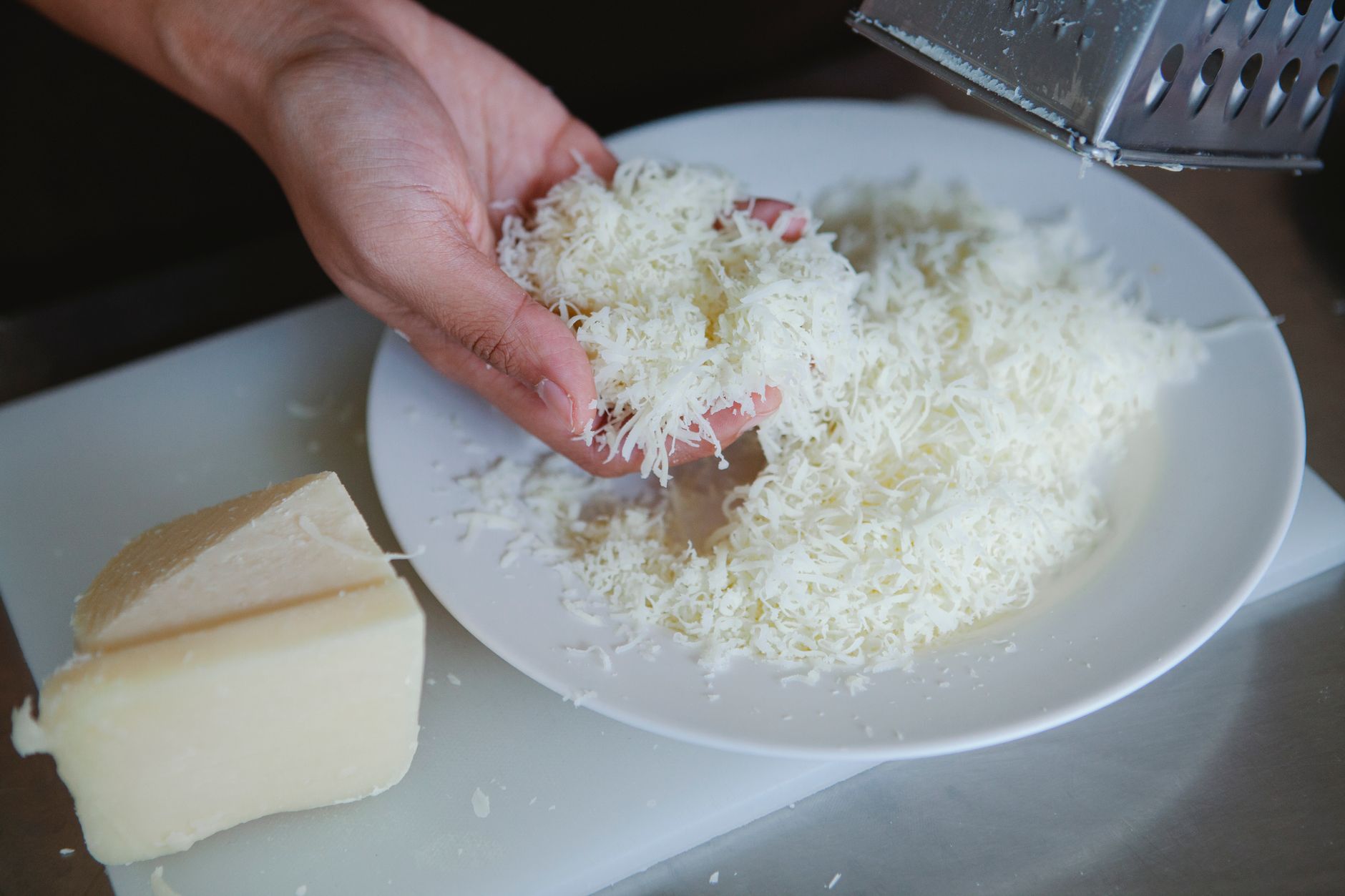 Mano toma queso blanco duro rallado de un plato, junto a un trozo de queso y un rallador metálico